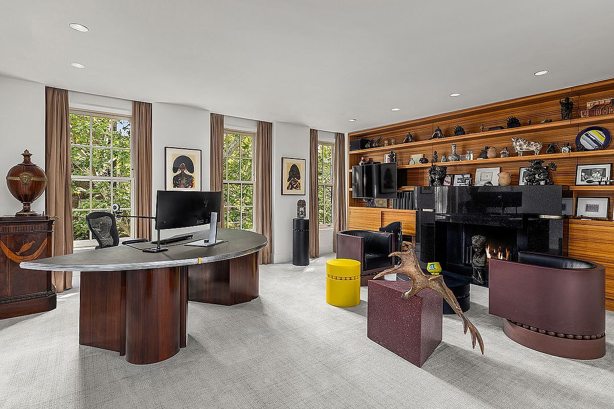 This sophisticated home office features a large, curved desk as the centerpiece, positioned in front of tall windows that allow for ample natural light. A prominent black stone fireplace surround serves as a focal point, flanked by extensive wood shelving filled with decorative objects and art. The room is furnished with modern seating, including two dark leather armchairs and a vibrant yellow stool, creating a professional yet artistic atmosphere.