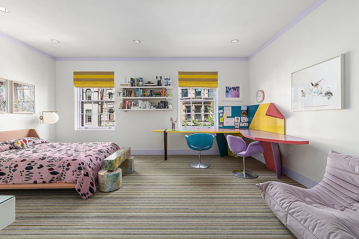 This vibrant and playful bedroom features a bed with a patterned pink duvet, a unique multi-colored desk setup, and a comfortable lounge chair. The room is accented by striped carpeting, yellow window treatments, and open shelving, creating a whimsical and creative atmosphere. The perspective is a wide-angle shot capturing the entire room, highlighting the colorful furniture and functional layout.