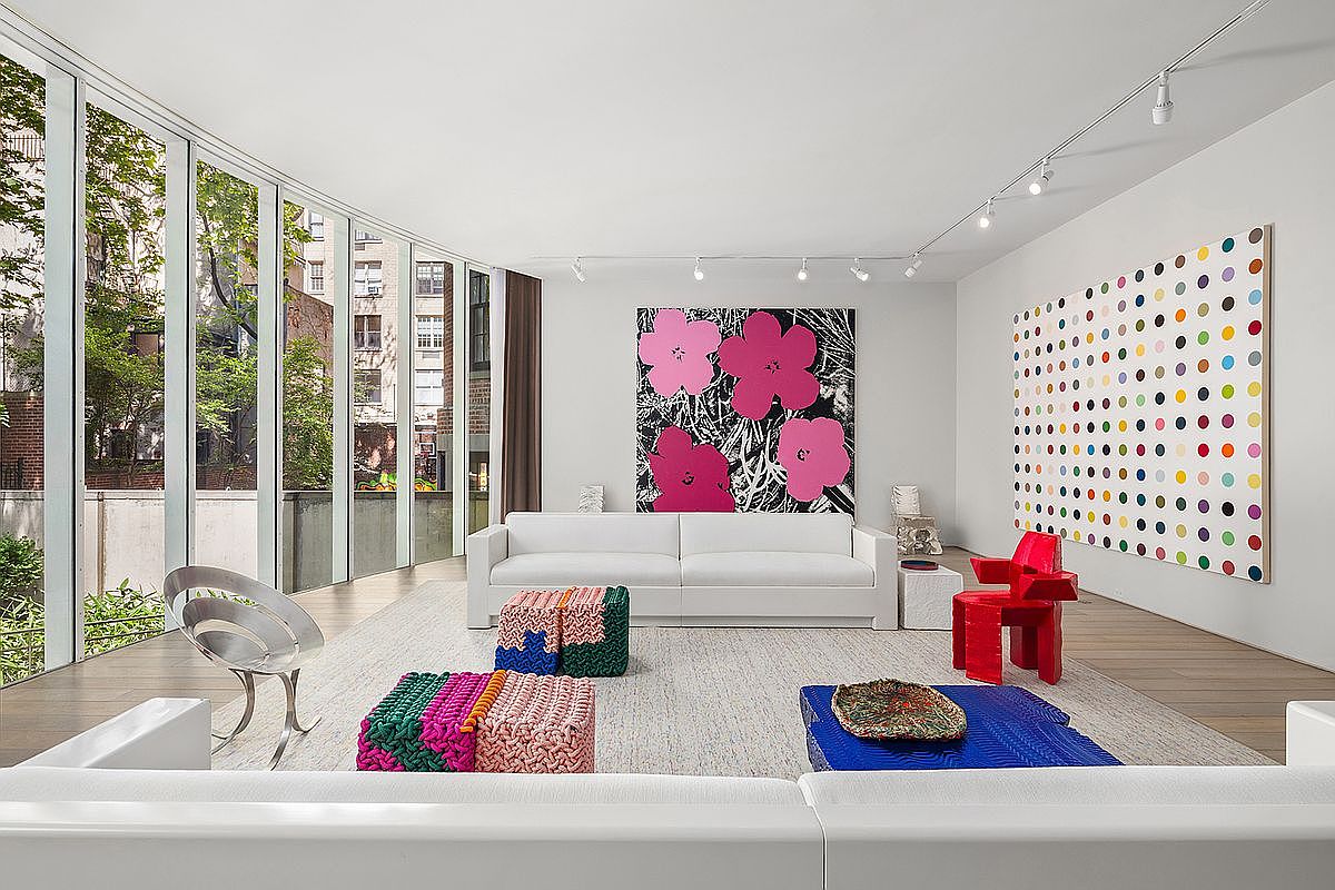 This expansive, modern living room is defined by a wall of floor-to-ceiling windows that provide an abundance of natural light and views of the surrounding greenery. The space features a minimalist white sofa, vibrant, textured ottomans, and a bold red sculptural chair, all set against a backdrop of large-scale contemporary art. The light wood flooring and clean, gallery-like white walls create a sophisticated, airy atmosphere perfect for showcasing high-end design.