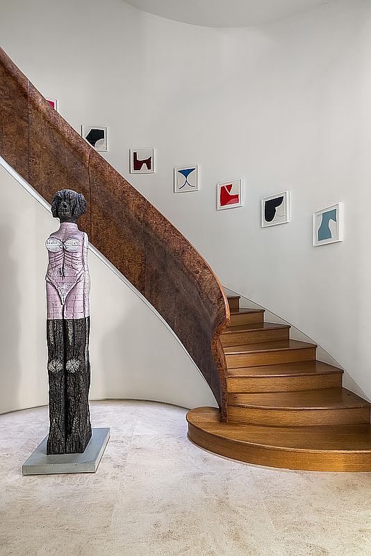 This elegant interior space features a striking curved staircase with warm-toned wooden treads and a distinctive, textured wood-paneled banister. A tall, abstract sculpture stands prominently on the light-colored stone floor at the base of the stairs, while a series of framed minimalist artworks adorn the curved white wall along the ascent. The perspective captures the graceful flow of the staircase, highlighting the sophisticated blend of artistic decor and architectural design.