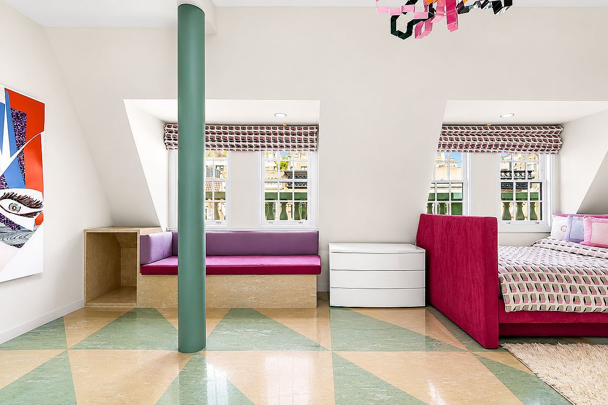 This vibrant bedroom features a playful, modern aesthetic with a geometric patterned floor and a bold magenta upholstered bed frame. A built-in window seat with purple cushions sits beneath two dormer windows, while a central green pillar adds a unique architectural element to the room. The space is bright and whimsical, accented by a colorful hanging light fixture and contemporary wall art.
