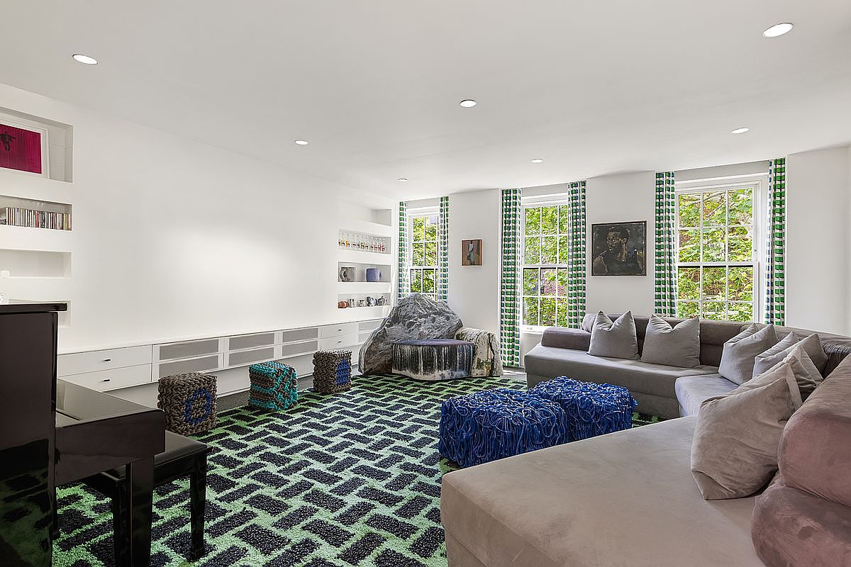 This spacious living room features a bold, geometric green and black patterned carpet that serves as the focal point of the design. The room is furnished with a large, plush grey sectional sofa, several textured blue cube ottomans, and a unique, rock-shaped accent chair. Built-in white shelving units line the wall, providing ample display space, while large windows with patterned curtains allow natural light to brighten the interior.