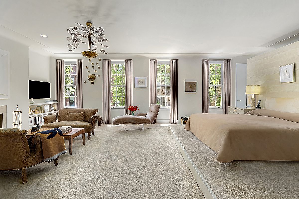 This spacious primary bedroom features a raised platform bed area, neutral carpeting, and a sophisticated seating area with a chaise lounge and a plush sofa. Three tall windows with elegant drapery allow natural light to fill the room, while a unique, multi-armed chandelier serves as a focal point above the seating area. The room is decorated with a minimalist aesthetic, including a wall-mounted television and subtle wall art, creating a serene and luxurious atmosphere.