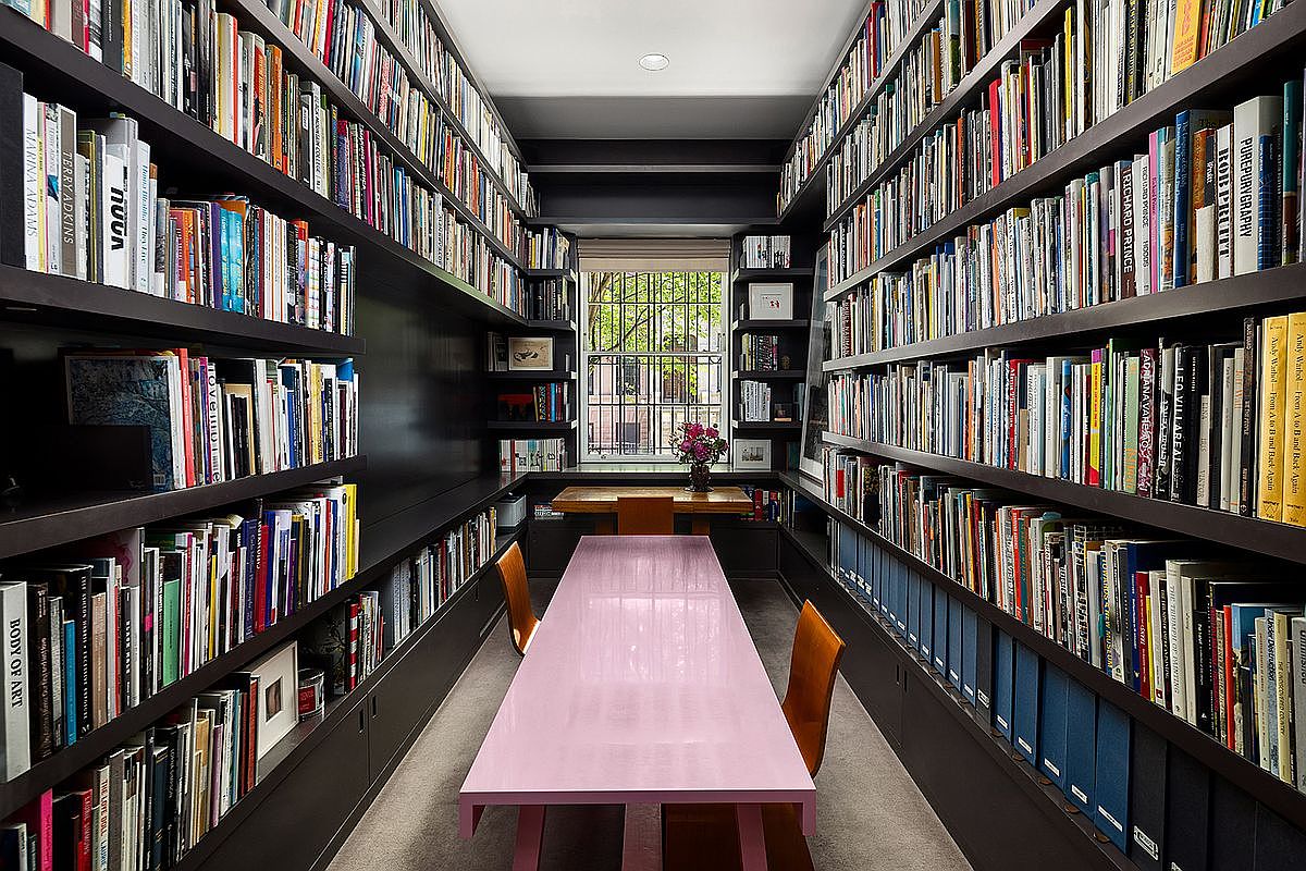 This sophisticated home library or study features dark, built-in shelving that spans both sides of the room, packed with an extensive collection of books. A long, light-colored table sits in the center, flanked by two wooden chairs, creating a focused workspace. The room is symmetrical and leads to a window at the far end, which provides natural light and a view of the exterior greenery.