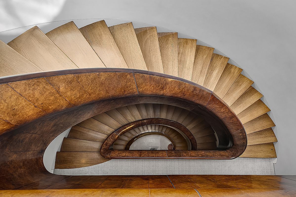 This image captures a striking, sculptural spiral staircase viewed from directly above, emphasizing its elegant, curved geometry. The design features light wood treads that contrast with a rich, dark wood-toned curved handrail and balustrade, creating a sophisticated and artistic focal point. The perspective highlights the rhythmic repetition of the steps as they descend into the center of the spiral, showcasing high-end craftsmanship and architectural detail.