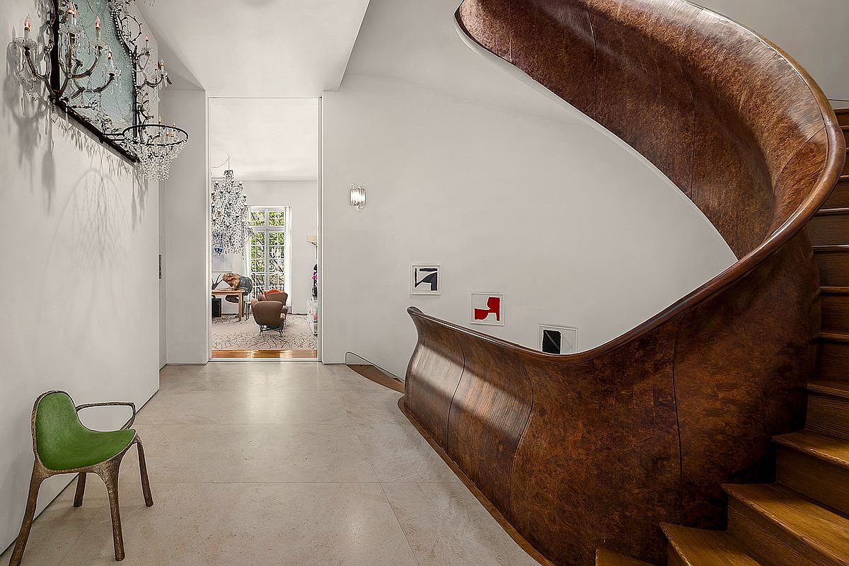This elegant entryway features a striking, curved staircase with a rich, wood-paneled balustrade that serves as a dramatic architectural focal point. The space is finished with light-toned stone flooring and minimalist white walls, accented by a unique green chair and modern art pieces. A view through the open doorway reveals a bright, secondary living space, creating a sense of depth and sophisticated flow throughout the home.