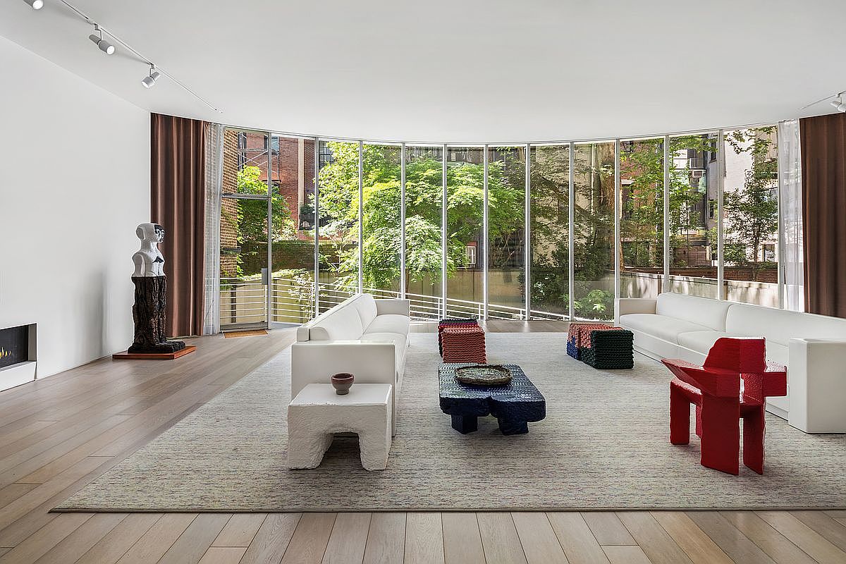 This expansive, modern living room features a curved wall of floor-to-ceiling windows that provide an abundance of natural light and views of the lush greenery outside. The space is anchored by a large, light-toned area rug, two white sofas, and a collection of sculptural, textured furniture pieces in bold colors. The light wood flooring and minimalist white walls create a bright, airy atmosphere, while a unique abstract sculpture stands near the corner.