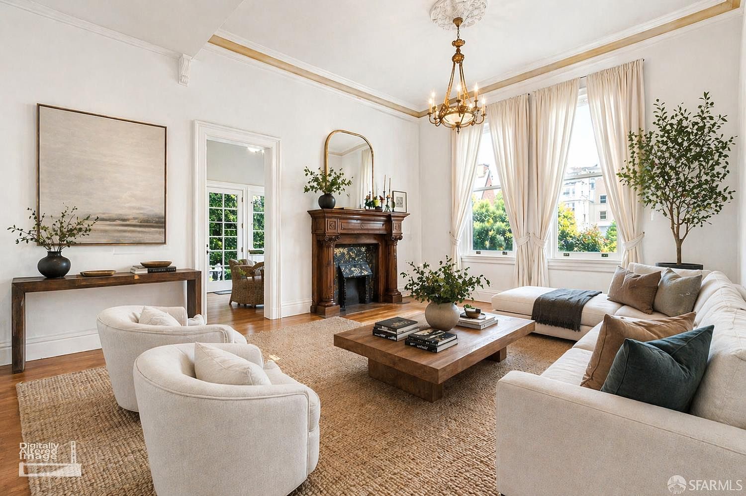 This elegant living room features a neutral color palette, a large wooden coffee table, and comfortable cream-colored seating arranged around a central rug. A prominent wood mantelpiece serves as a focal point, complemented by a tall mirror and a classic chandelier overhead. Large windows draped in soft curtains allow natural light to fill the space, creating an inviting and sophisticated atmosphere.