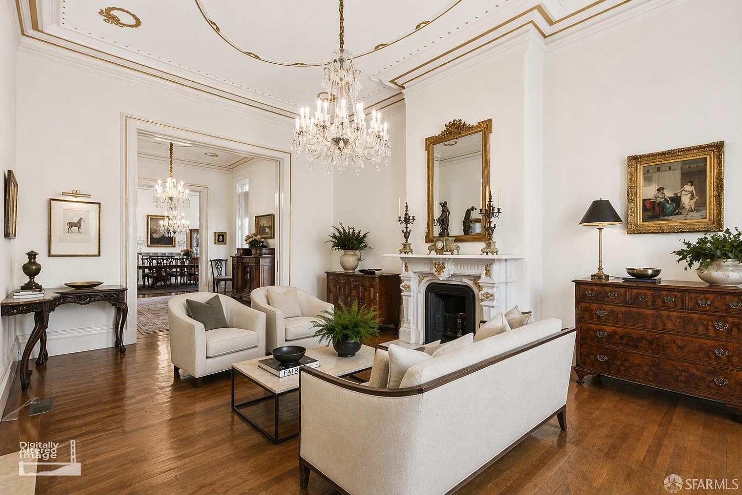 This elegant living room features a sophisticated, traditional aesthetic with high ceilings, intricate crown molding, and polished hardwood floors. The space is anchored by a white fireplace mantel with a large ornate mirror above, flanked by two comfortable armchairs and a plush sofa. A crystal chandelier serves as a focal point, while a view into the adjacent dining room highlights the home's open, flowing floor plan.