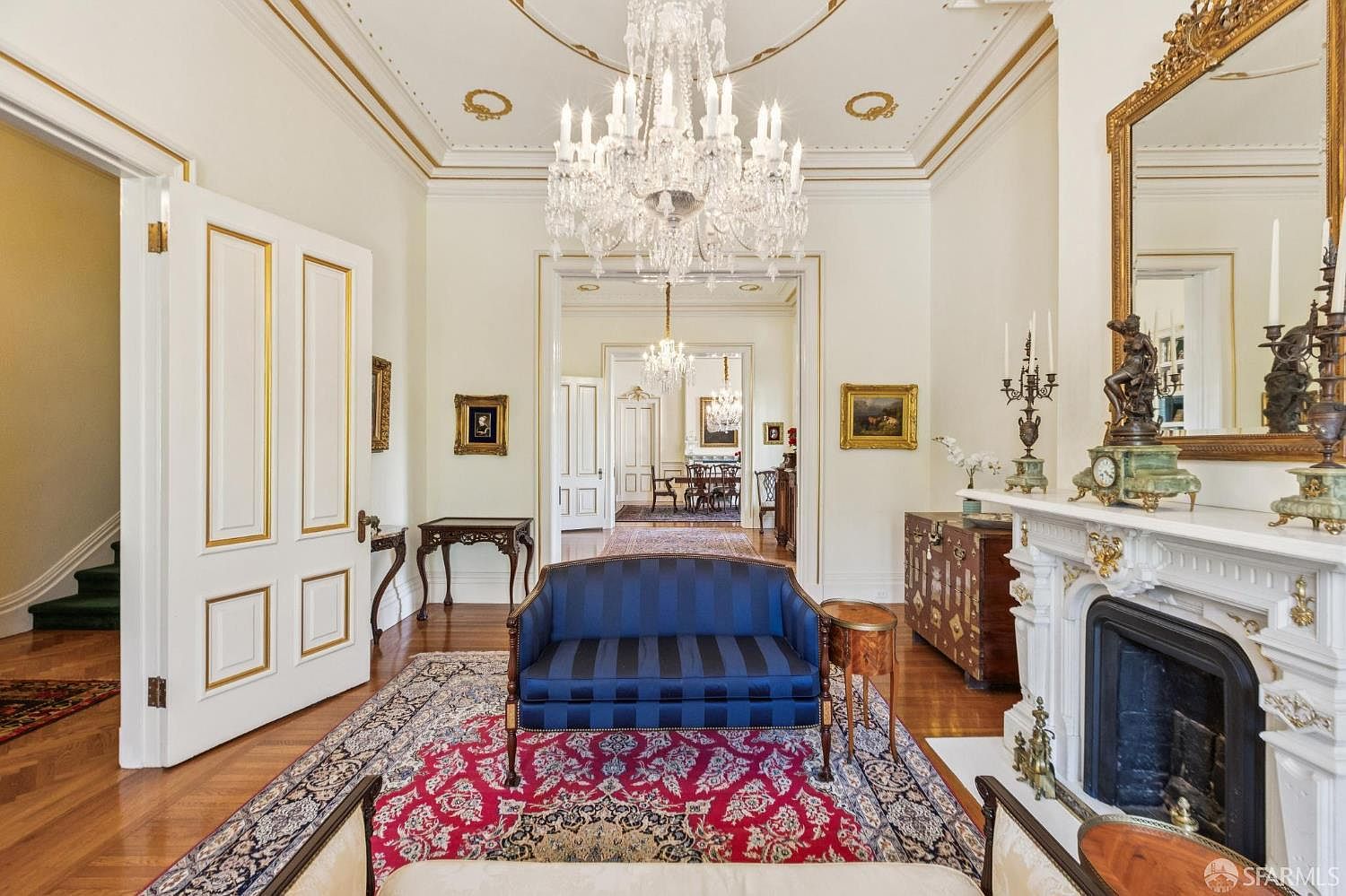 This elegant formal living room features a classic aesthetic with a prominent crystal chandelier, ornate crown molding, and a decorative fireplace with a detailed mantel. A blue striped settee sits atop a vibrant red patterned rug, while a large mirror hangs above the fireplace, reflecting the sophisticated space. The room opens into a series of connected rooms, showcasing a grand, traditional architectural layout with polished wood floors.