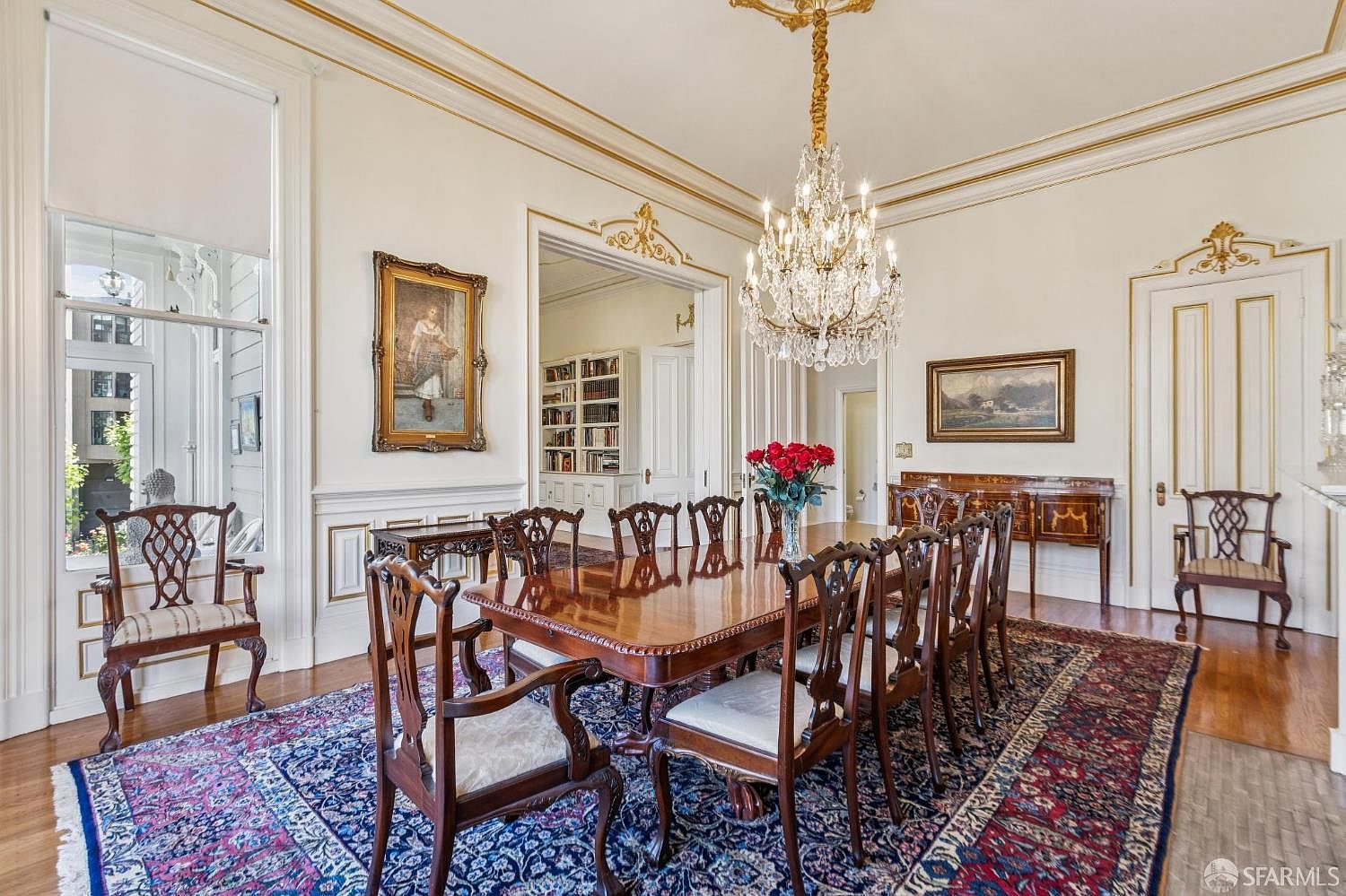 This elegant formal dining room features a large, polished wood table surrounded by classic carved chairs, set atop a rich, intricately patterned area rug. A grand crystal chandelier serves as the focal point, suspended from a high ceiling with decorative molding, while a sideboard sits against the wall beneath a landscape painting. The space is framed by tall windows and a wide doorway leading to an adjacent library, creating a sophisticated and traditional atmosphere.