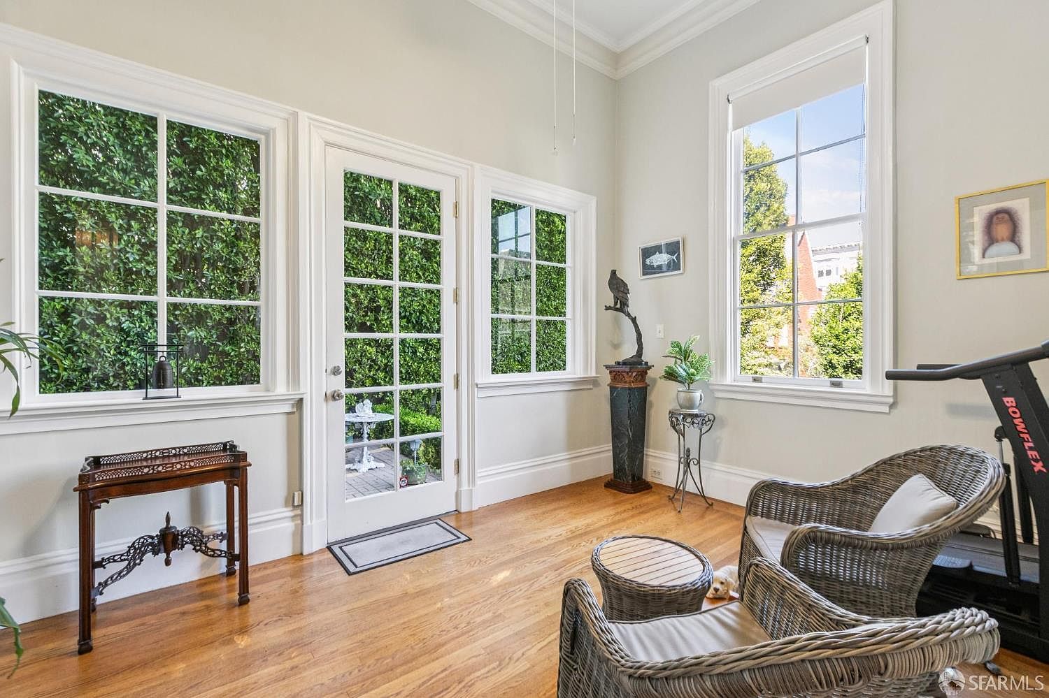 This bright sunroom features light-toned walls, warm wood flooring, and multiple large windows that provide ample natural light and views of the lush greenery outside. The space is furnished with wicker armchairs, a small side table, and a pedestal displaying a sculpture, creating a relaxed and inviting atmosphere. A glass-paned door leads directly to an outdoor patio, seamlessly blending indoor and outdoor living spaces.