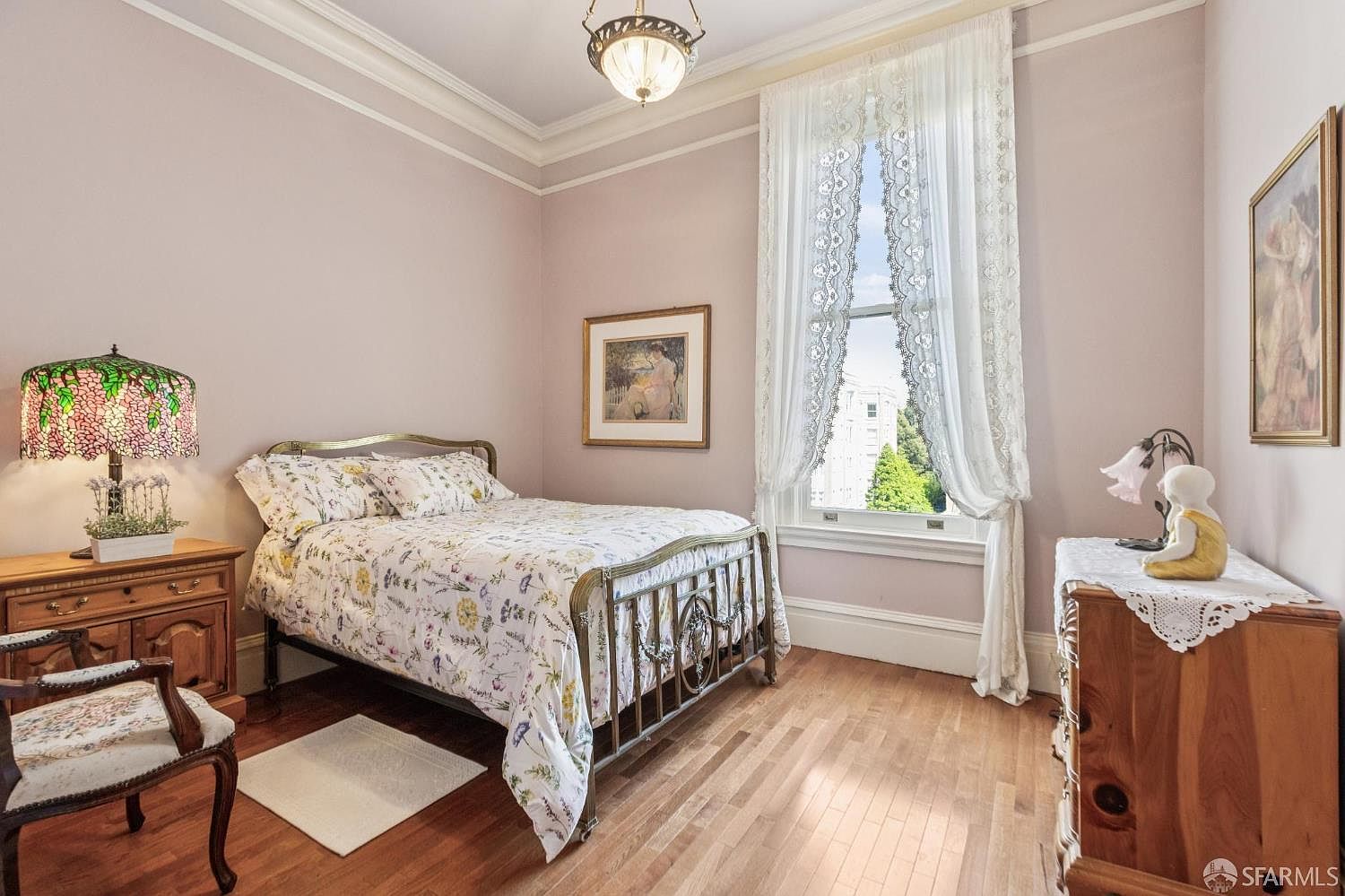 This charming guest bedroom features light-toned walls, hardwood flooring, and a metal-framed bed dressed in floral bedding. A large window with delicate lace curtains allows natural light to fill the room, while a vintage-style lamp and decorative artwork add a touch of classic elegance. The space is furnished with a wooden dresser and a patterned armchair, creating a cozy and inviting atmosphere.