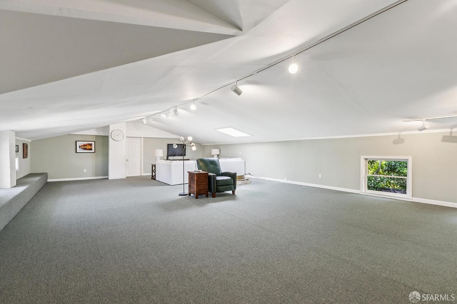 This spacious attic room features a dramatic sloped ceiling and neutral carpeting throughout. The area is furnished with a comfortable green armchair, a small wooden side table, and a white sofa set positioned near a television. A window on the right provides natural light, while track lighting mounted on the ceiling illuminates the expansive, open-concept living space.
