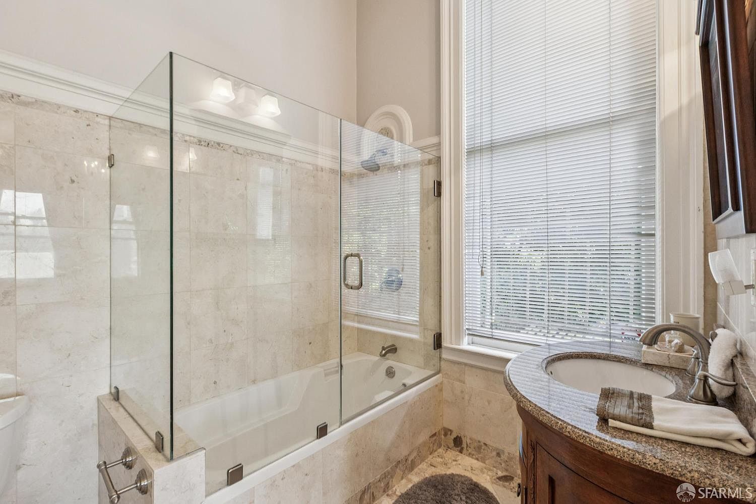 This bathroom features a combination tub and shower enclosed by a glass panel, finished with light-toned stone tile walls. A curved vanity with a stone countertop and an undermount sink sits beside a window covered by horizontal blinds. The space is bright and neutral, offering a functional and clean aesthetic.