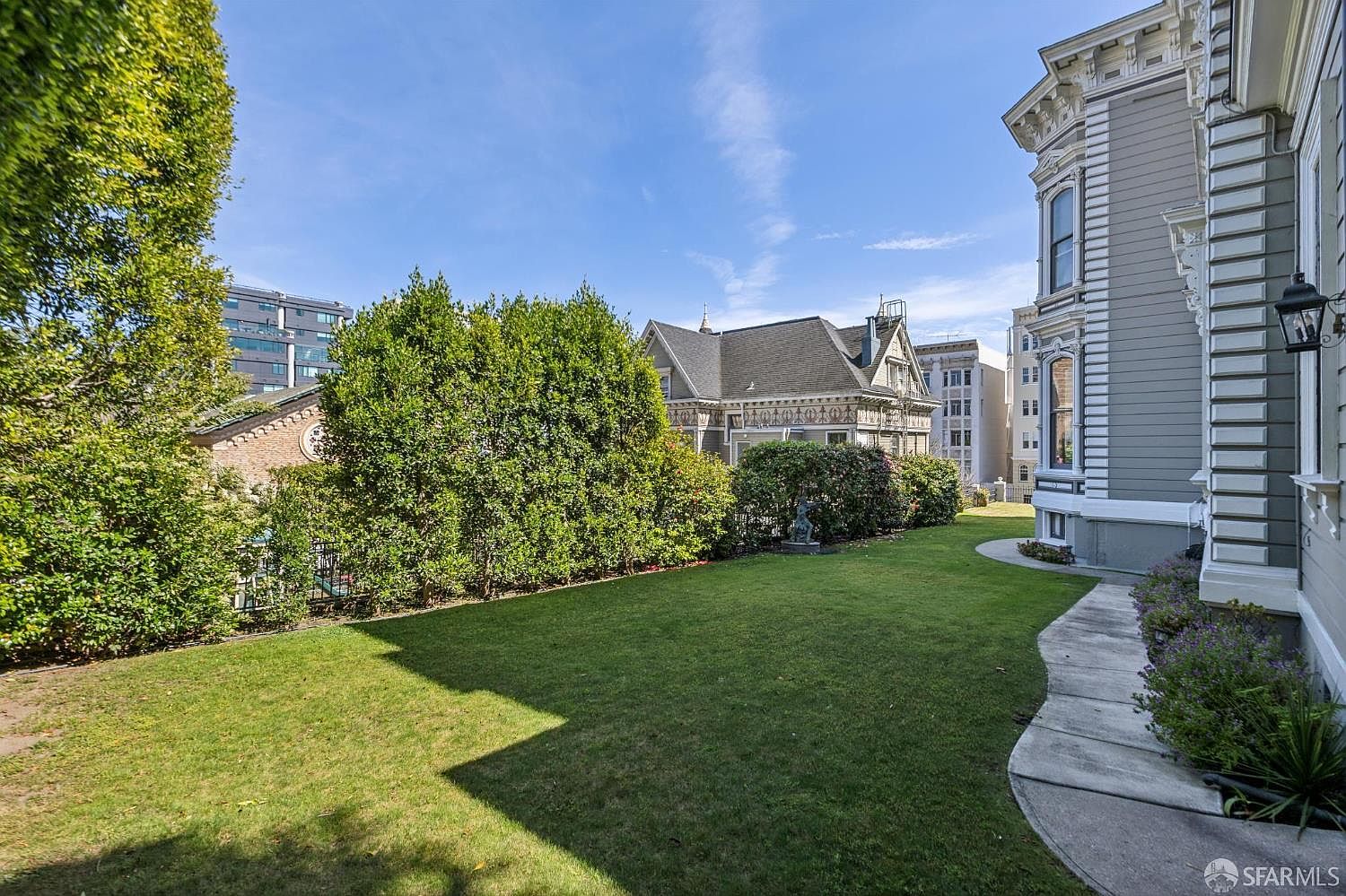 This side yard features a well-manicured lawn bordered by a lush, mature hedge that provides privacy. A concrete pathway runs alongside the side of a classic, multi-story Victorian-style home with grey siding and intricate architectural detailing. The perspective captures the transition from the home's exterior to the open green space, highlighting the property's peaceful outdoor setting.