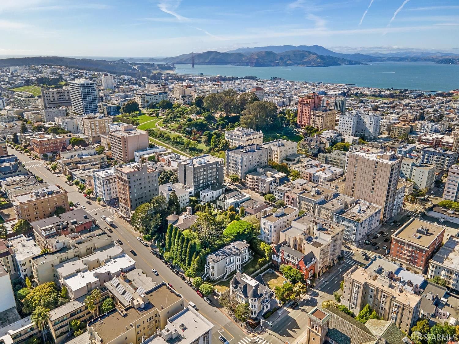 This high-angle aerial view captures a dense urban neighborhood in San Francisco, showcasing a mix of historic Victorian architecture and modern high-rise apartment buildings. The perspective looks out toward the iconic Golden Gate Bridge and the bay, with a lush green park nestled in the center of the cityscape. The scene conveys the vibrant, hilly character of the city with its intricate grid of streets and diverse building styles.