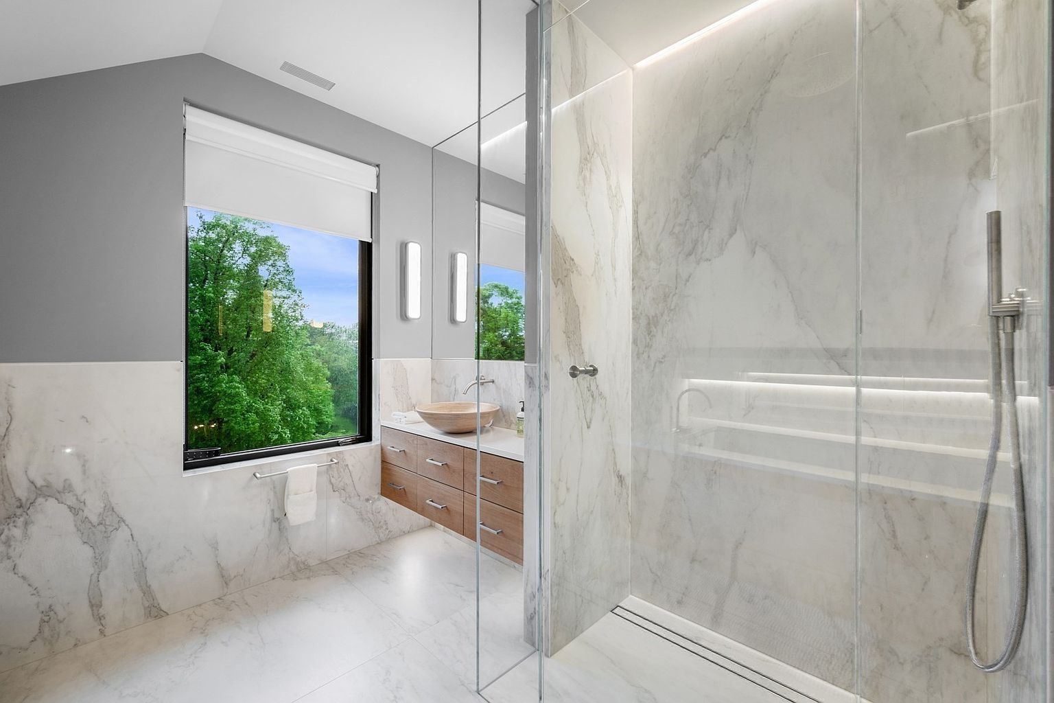 This modern primary bathroom features a sleek, glass-enclosed walk-in shower with light-toned stone wall cladding and integrated lighting. A floating vanity with light wood cabinetry and a stone vessel sink sits beneath a large mirror, while a window provides a bright, scenic view of the surrounding trees. The space is finished with light-colored stone flooring, creating a clean, spa-like atmosphere.