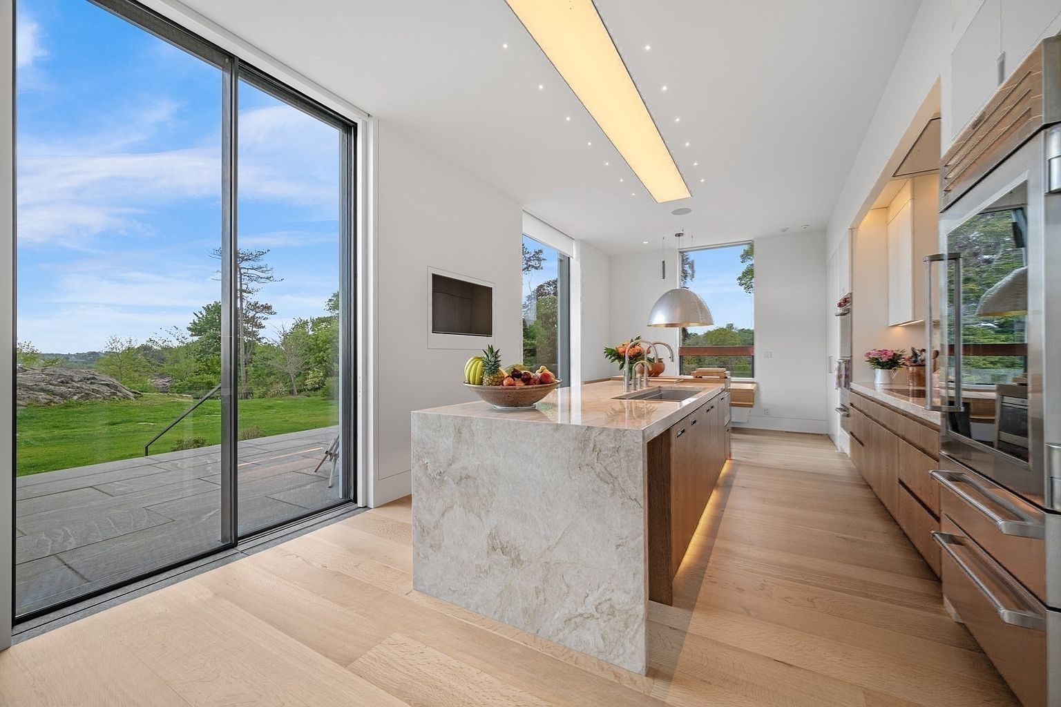 This modern, minimalist kitchen features a large central island with a stone waterfall edge and light wood cabinetry. The space is bathed in natural light from expansive sliding glass doors that open to a stone patio and a lush green landscape. A sleek, linear light fixture hangs above the island, complementing the clean lines and neutral color palette of the room.