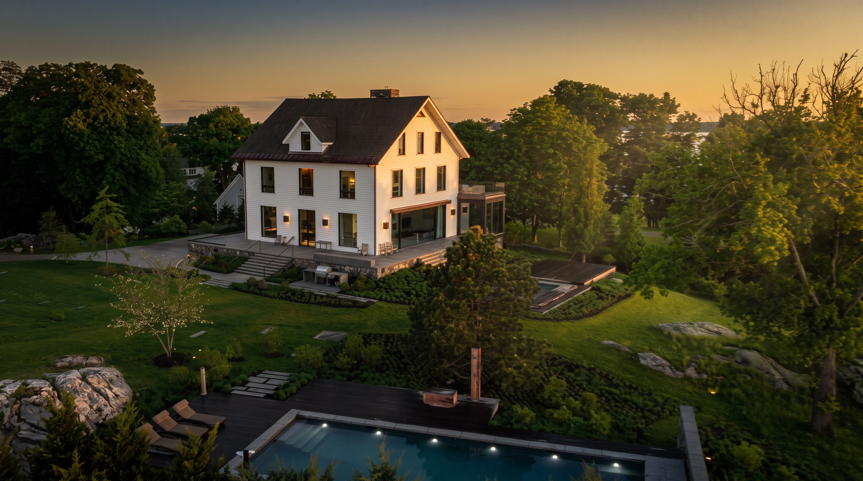 This aerial perspective captures a stately white home set within a lush, expansive landscape featuring mature trees and rocky outcrops. The property includes a rectangular swimming pool with a dark wood deck, a stone-paved patio area, and a modern glass-enclosed addition extending from the main structure. The scene is bathed in the soft, golden light of dusk, highlighting the elegant architecture and the serene, private outdoor living spaces.