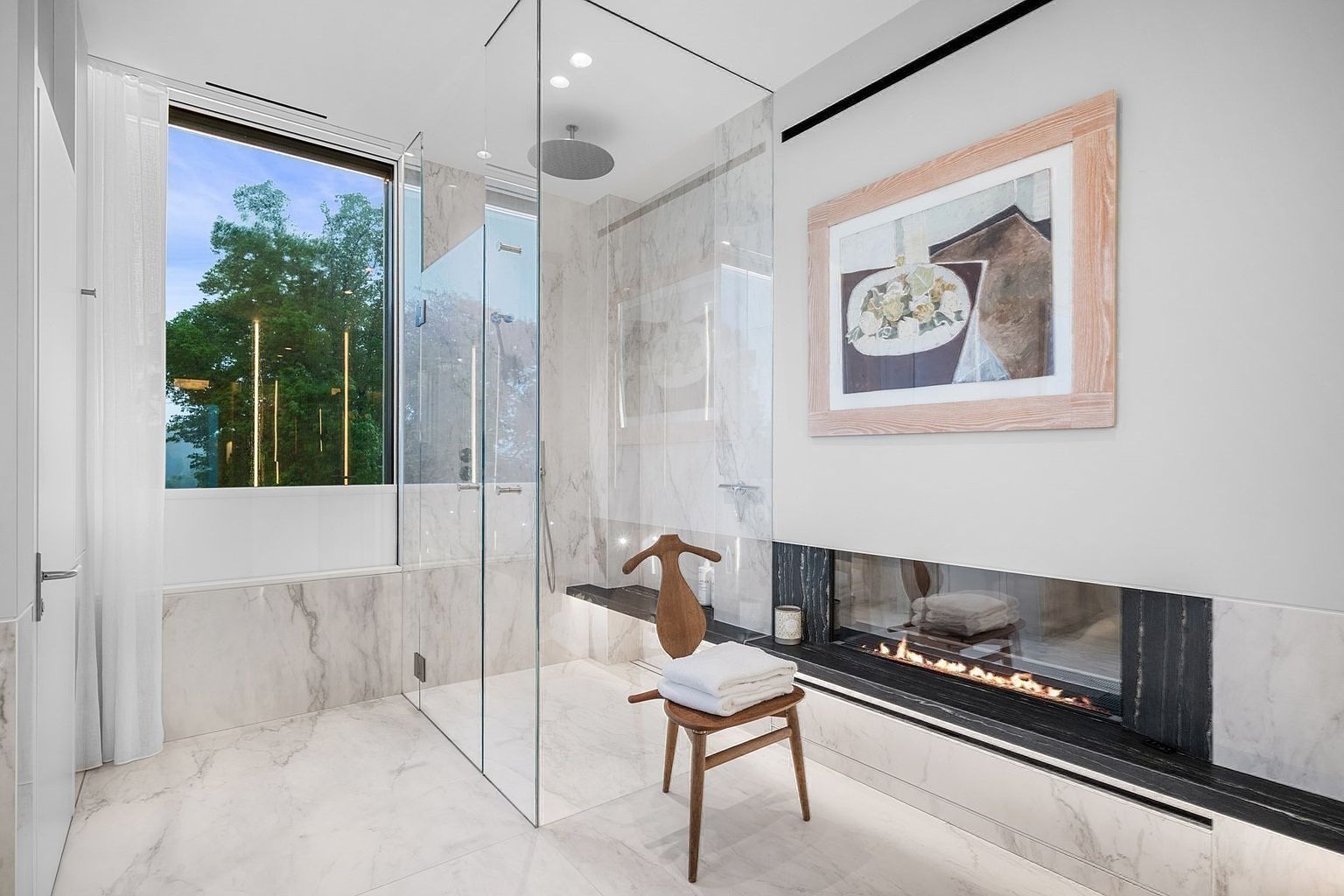 This luxurious primary bathroom features a glass-enclosed walk-in shower with a rainfall showerhead and a built-in stone bench. A linear fireplace is integrated into the wall, adding warmth and a spa-like atmosphere, while a wooden chair with a stack of white towels sits in the foreground. The space is finished with light-toned stone surfaces and large windows that provide a view of the surrounding greenery.