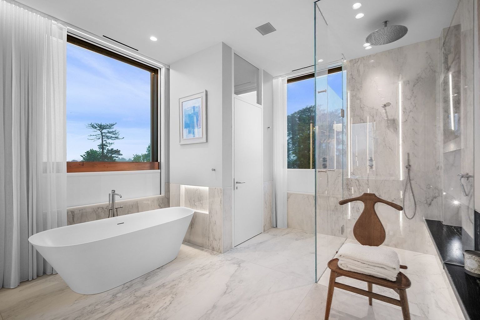 This luxurious primary bathroom features a sleek, freestanding soaking tub positioned beneath a large window that offers expansive outdoor views. To the right, a spacious walk-in shower is enclosed by glass panels and finished with light-toned stone wall cladding. A wooden stool with a towel sits in the foreground, adding a warm, organic touch to the bright, minimalist aesthetic of the space.
