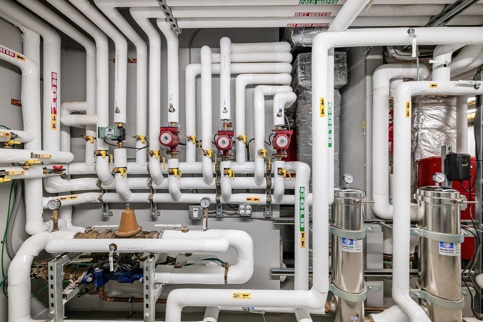 This image displays a complex mechanical room featuring a dense network of white-insulated pipes, multiple red circulation pumps, and industrial filtration units. The setup is highly organized, showcasing professional-grade plumbing and HVAC infrastructure typical of a large residential or commercial building's utility area. The perspective is a direct, eye-level view that emphasizes the intricate layout and technical maintenance of the property's core systems.