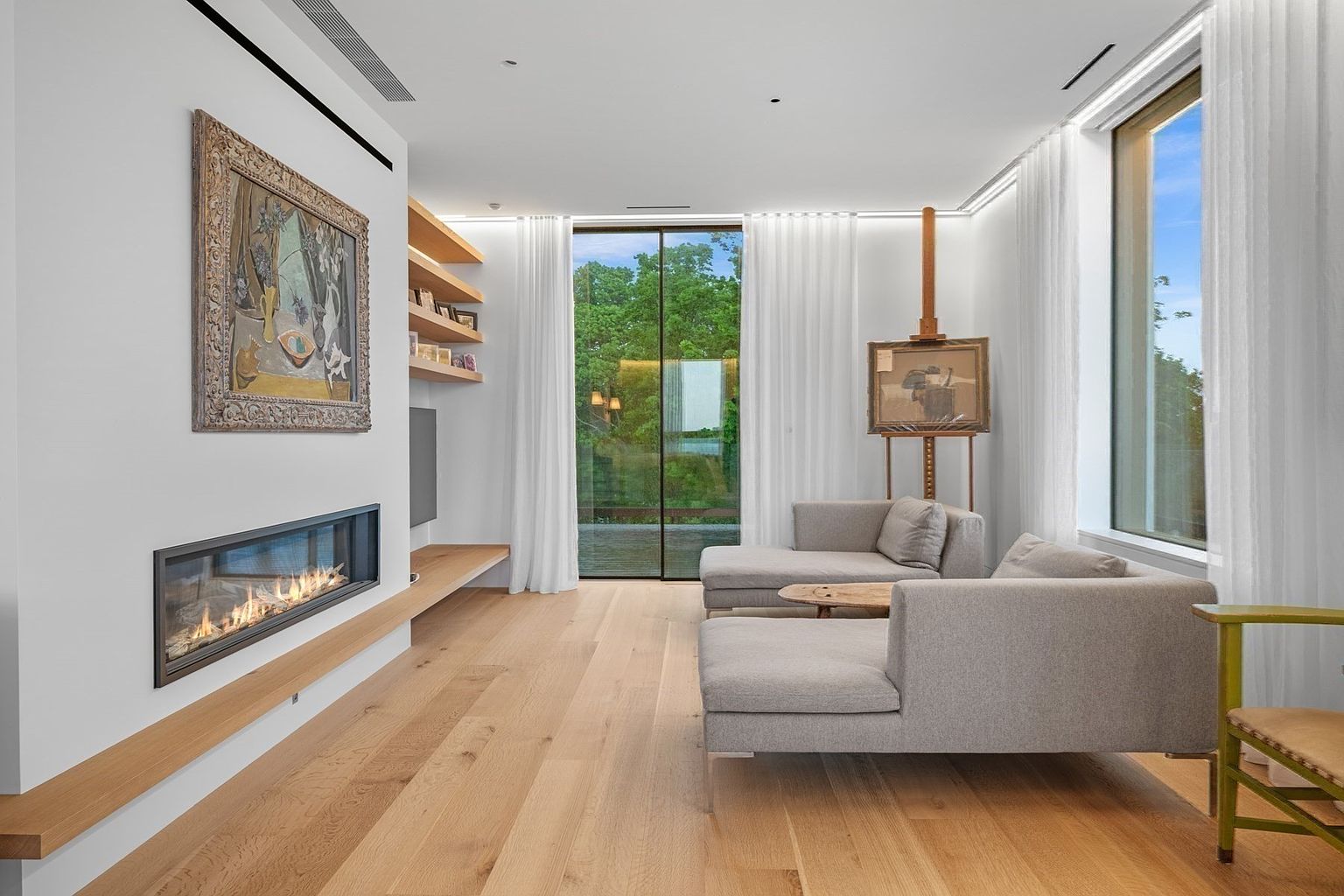 This bright and airy living room features light wood flooring and a minimalist aesthetic, anchored by a sleek, linear fireplace set into a white wall. A pair of grey sectional sofas are arranged around a small wooden coffee table, while an artist's easel stands near large windows that provide a view of the lush greenery outside. The space is accented by built-in wooden shelving and a framed painting, creating a sophisticated and tranquil atmosphere.