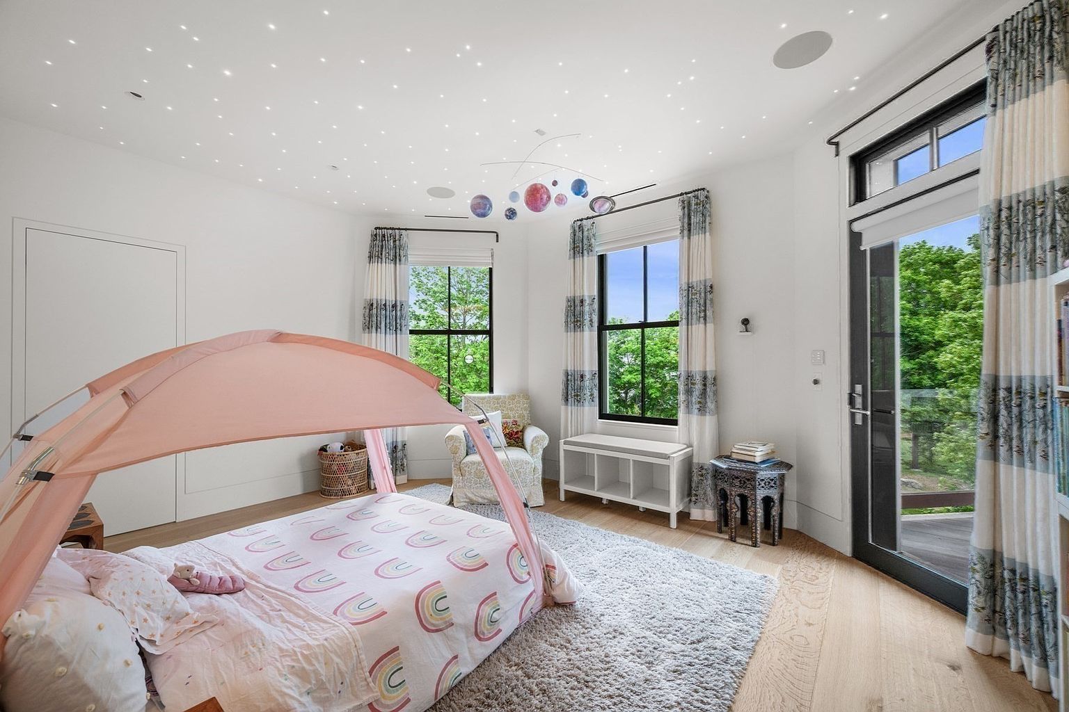 This charming child's bedroom features a whimsical canopy bed with a rainbow-patterned duvet and a unique ceiling treatment designed to mimic a starry night sky. The room is bright and airy, with light wood flooring, a cozy reading nook with an armchair, and large windows that provide ample natural light and a view of the surrounding greenery. A mobile of planets hangs from the ceiling, adding a playful, educational touch to the space.