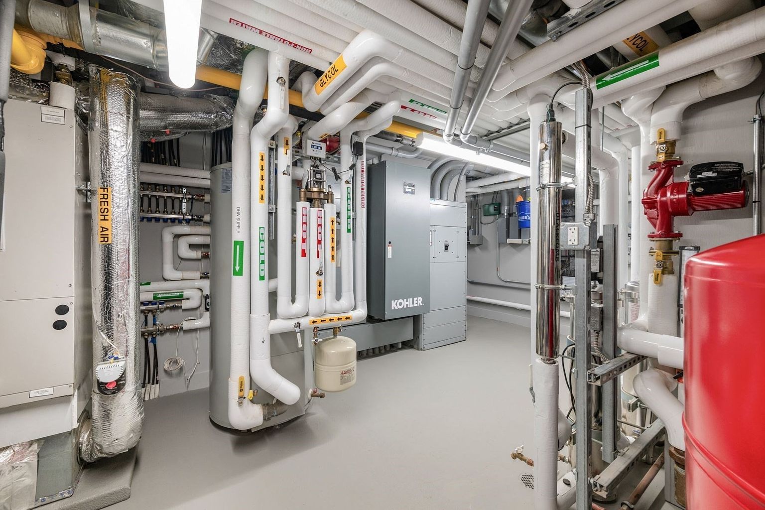 This image shows a mechanical room featuring a complex network of industrial pipes, valves, and HVAC equipment. A large water heater and a Kohler electrical panel are prominent, set against a clean, utilitarian space with grey flooring. The perspective is a wide-angle view that captures the intricate infrastructure typical of a high-end residential or commercial building's utility area.