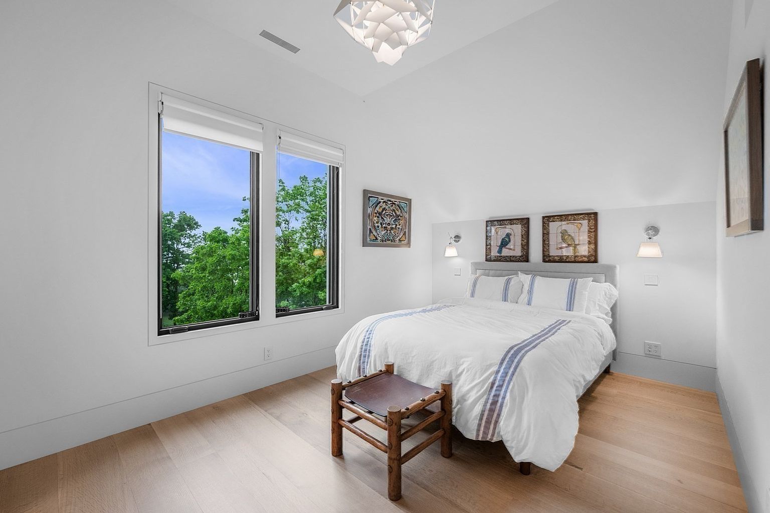 This bright and airy bedroom features light wood flooring and crisp white walls, creating a clean, minimalist aesthetic. A queen-sized bed with white bedding is centered against the wall, flanked by two wall-mounted sconces and framed bird artwork. Large windows on the left provide ample natural light and a view of the surrounding greenery, while a unique geometric pendant light hangs from the vaulted ceiling.
