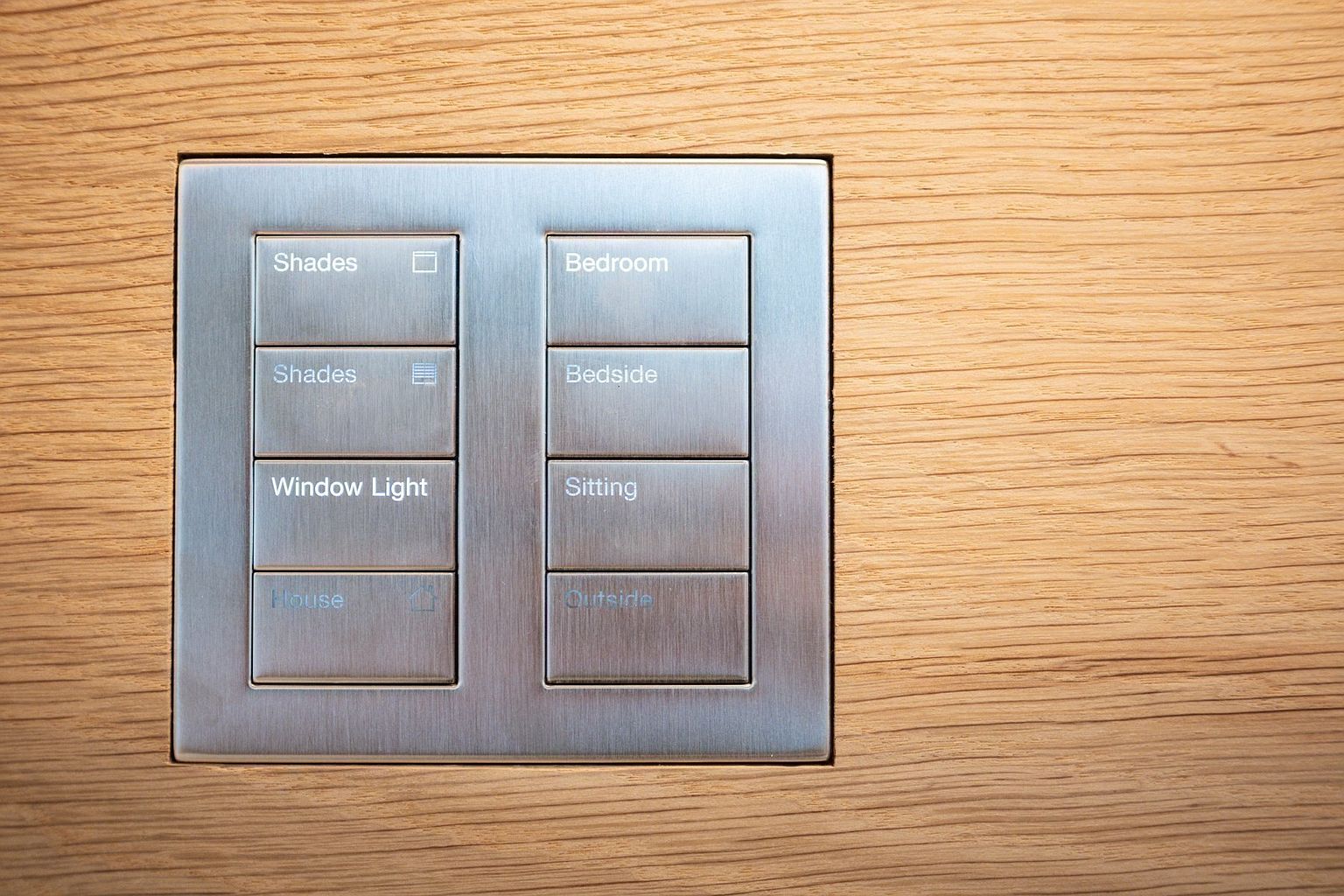 A close-up, eye-level shot of a modern, brushed-metal smart home control panel mounted flush into a light wood-paneled wall. The interface features two columns of backlit buttons with clear, minimalist labels for controlling various household functions like shades, lighting, and specific room zones. The clean, industrial aesthetic suggests a high-end, automated home environment.