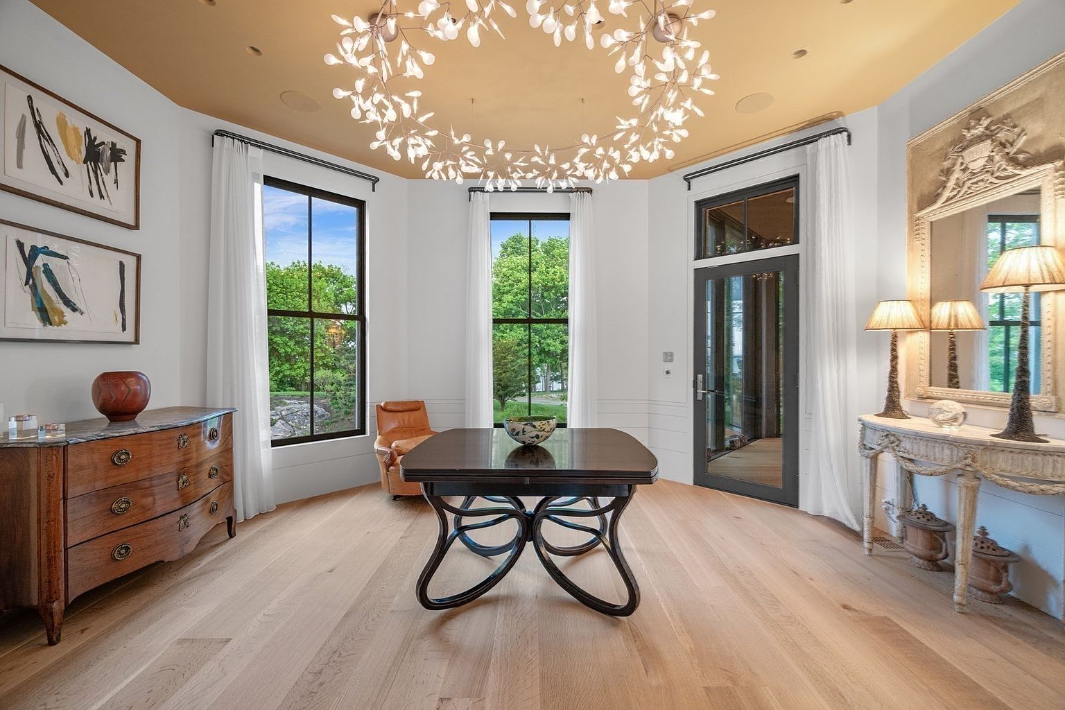 This elegant room features light wood flooring and a distinctive circular light fixture suspended from a warm-toned ceiling. A central dark wood table serves as a focal point, complemented by a classic wooden dresser on the left and a decorative console table with lamps on the right. Large windows provide ample natural light and views of the surrounding greenery, creating a bright and sophisticated atmosphere.