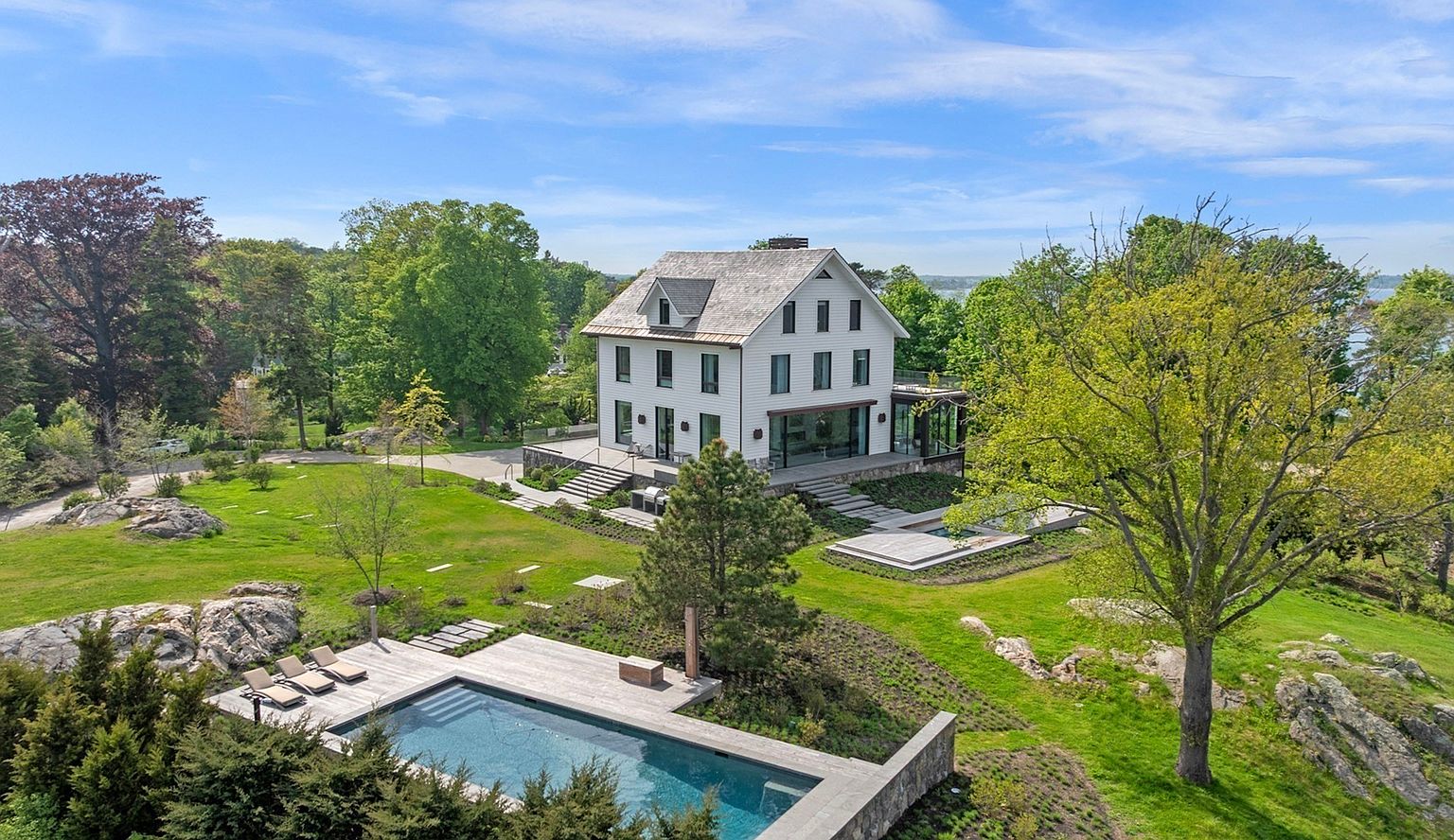 This high-angle aerial view captures a sophisticated, multi-story white residence set within a sprawling, lush landscape. The property features a modern swimming pool with a stone deck, expansive green lawns, and mature trees, all set against a backdrop of distant water. The home showcases a classic architectural silhouette with a shingled roof and large windows, emphasizing a seamless connection between the refined interior and the natural surroundings.