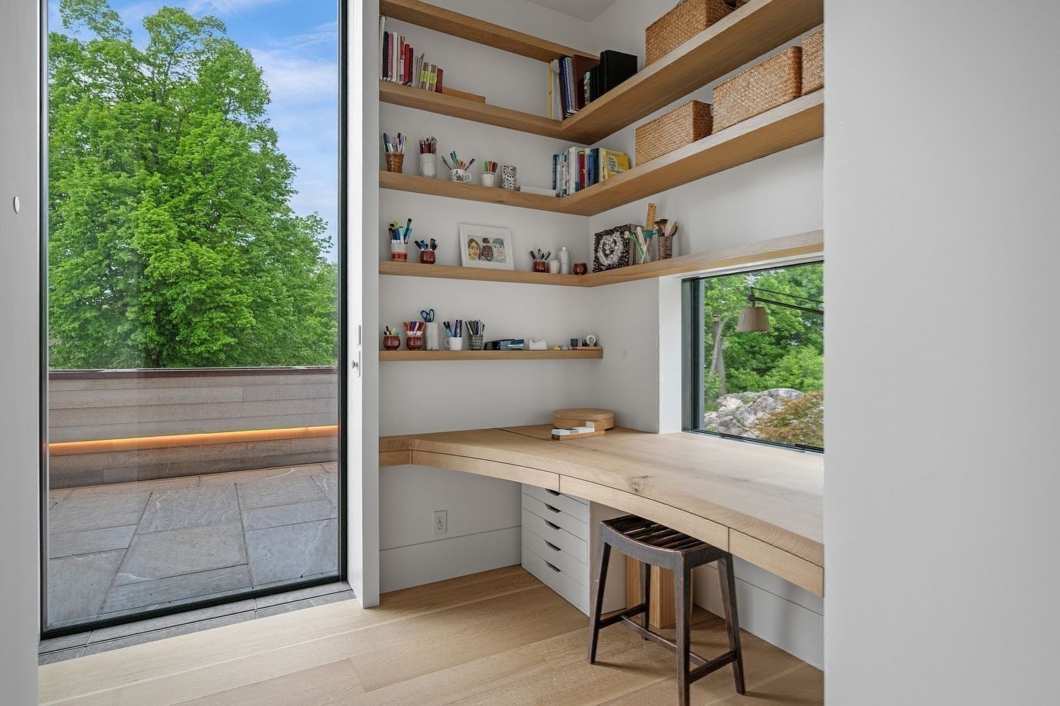 This modern home office features a custom light wood desk integrated into a corner nook, complemented by matching open shelving that provides ample storage for books and decor. A large window beside the desk offers a serene view of the outdoors, while a floor-to-ceiling glass door provides access to an adjacent balcony. The space is bright and minimalist, emphasizing clean lines and a seamless connection to the natural environment.