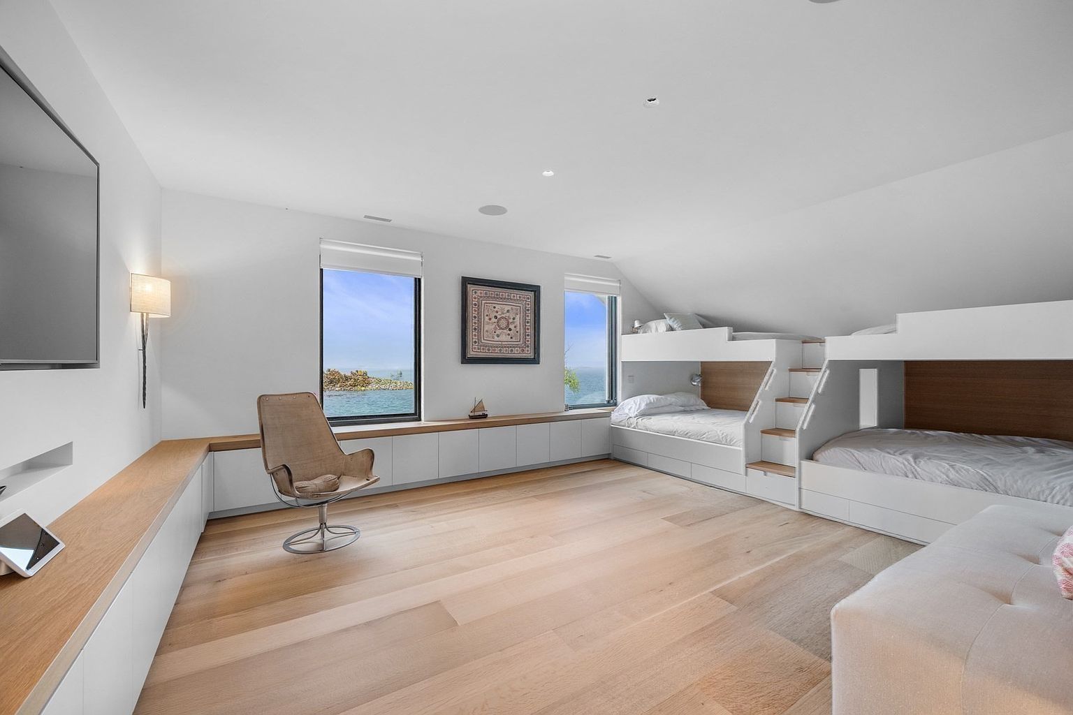 This bright and airy bedroom features a custom built-in bunk bed configuration with integrated stairs and light wood accents. The room is anchored by light wood flooring and a long, low-profile storage unit that runs beneath two windows overlooking the water. A modern, minimalist chair sits in the center of the room, which is finished with clean white walls and recessed lighting, creating a serene and functional space.