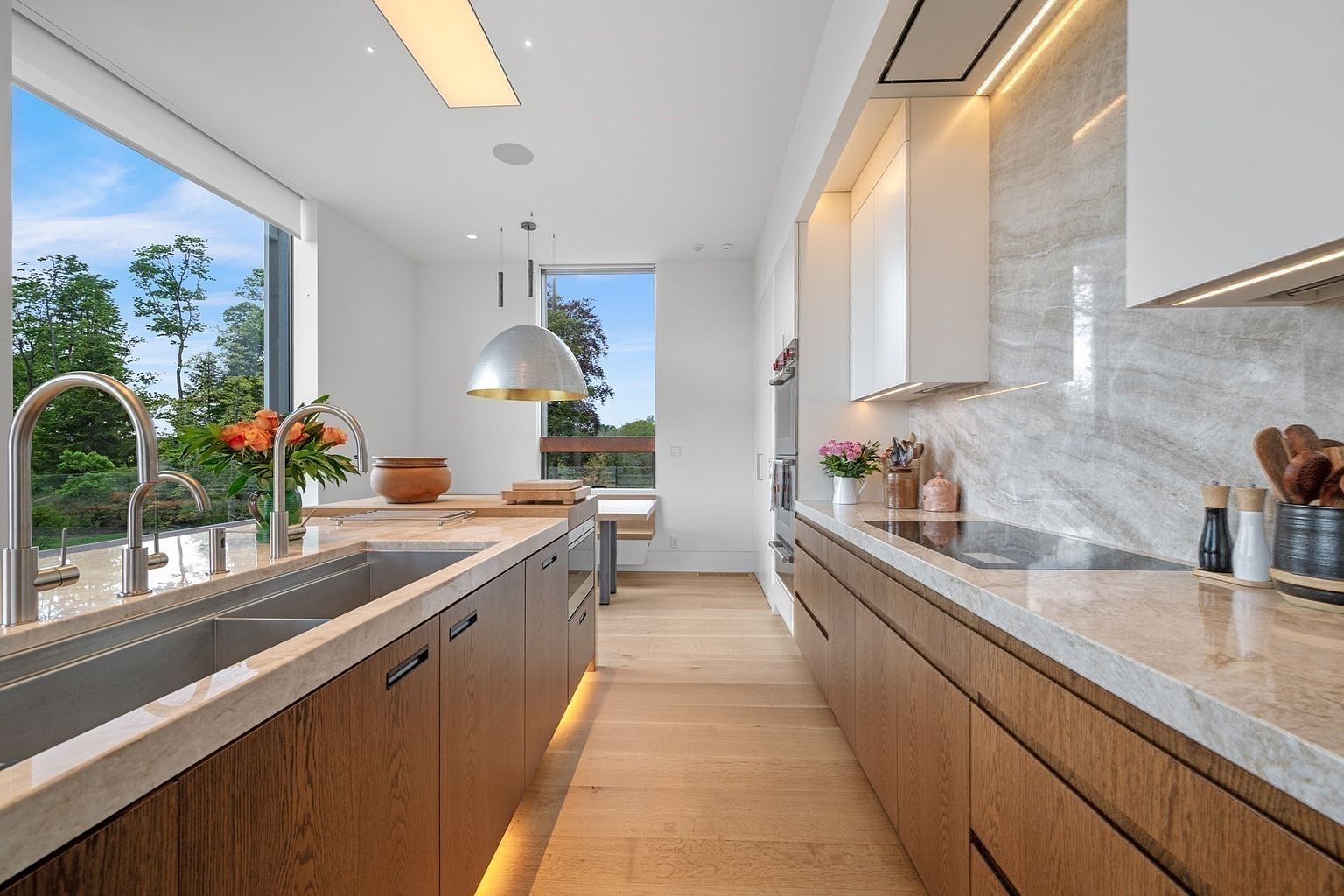 This modern kitchen features a sleek, linear layout with warm-toned wood cabinetry and light-colored stone countertops. A large island with a double sink serves as a central focal point, complemented by a large pendant light and expansive windows that provide a bright, airy atmosphere with views of the surrounding greenery. The space is finished with light wood flooring and integrated appliances, creating a clean, sophisticated aesthetic.