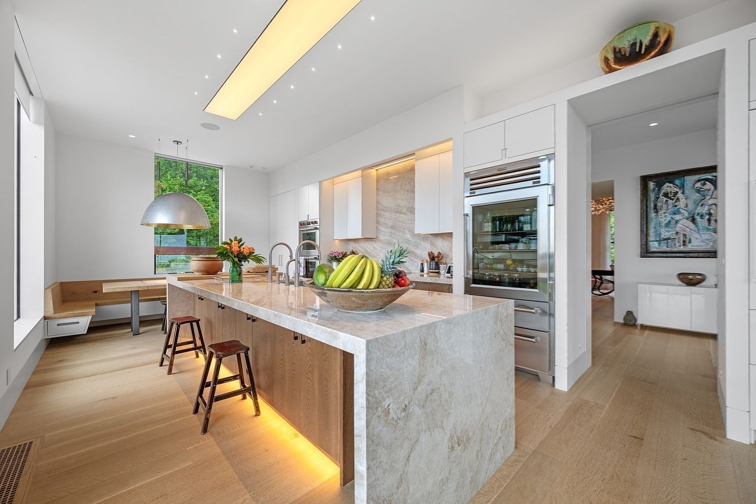 This modern kitchen features a large island with a stone countertop and light wood cabinetry, complemented by two wooden stools. The space is illuminated by a long, recessed light fixture and features a built-in stainless steel refrigerator and a cozy breakfast nook with bench seating by a large window. The overall aesthetic is bright and contemporary, with light wood flooring and a seamless transition into an adjacent hallway.