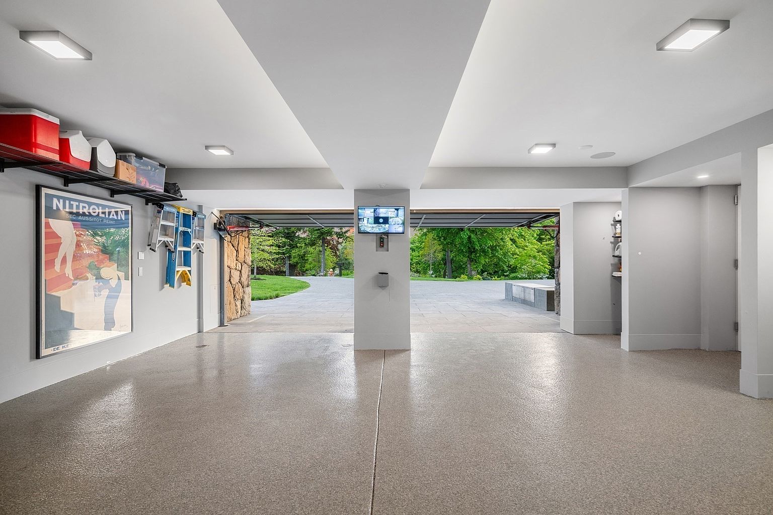 This spacious, well-lit garage features a clean, neutral aesthetic with a speckled epoxy floor and light-colored walls. The open garage doors reveal a paved driveway and a lush, green outdoor landscape, creating a seamless transition between the indoor storage space and the exterior grounds. A central pillar holds a small wall-mounted television, while a large vintage-style poster adds a decorative touch to the side wall.