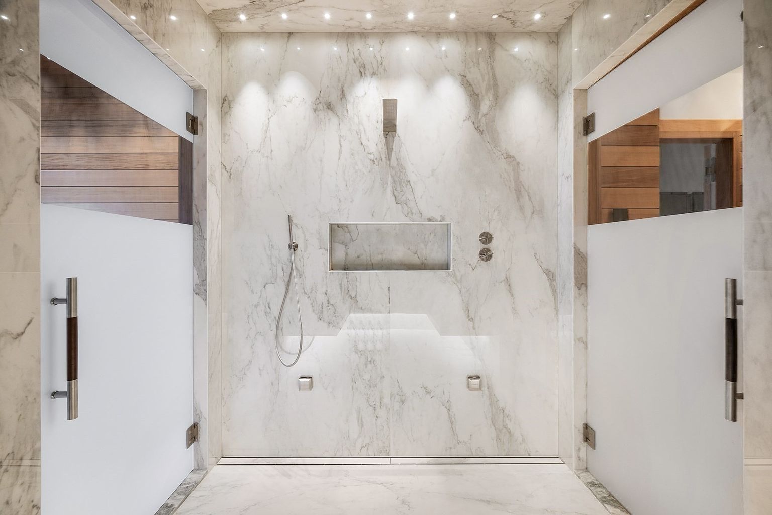 This luxurious walk-in shower features expansive stone wall cladding with prominent veining and recessed accent lighting. The space is equipped with a modern rainfall showerhead, a handheld sprayer, and a built-in wall niche for storage, all framed by frosted glass doors on either side. The minimalist design is enhanced by integrated floor-level lighting and a sleek, linear drain, creating a high-end, spa-like atmosphere.