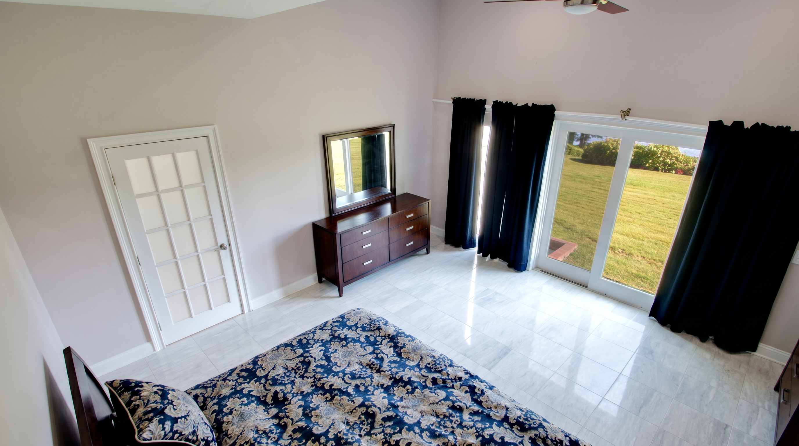 This spacious primary bedroom features high vaulted ceilings, elegant patterned blue and beige bedding, and glossy marble-tiled floors. The room is anchored by a dark wood dresser with a matching mirror and benefits from ample natural light through large glass sliding doors draped with stark black curtains. A classic white French door adds a touch of architectural charm to this clean, neutral-walled retreat.