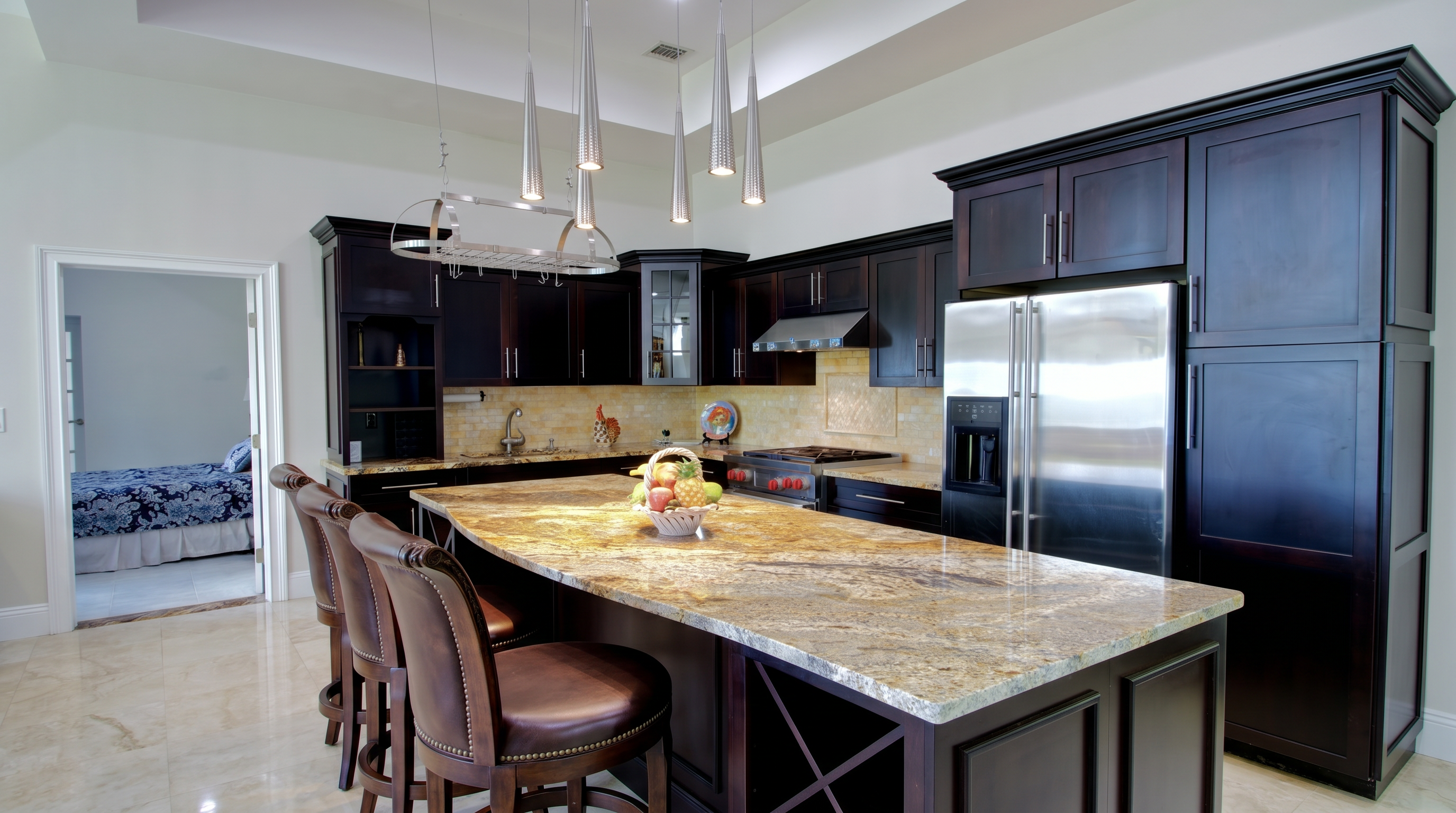 This sophisticated kitchen features dark wood cabinetry, a large granite-topped island with leather bar stools, and high-end stainless steel appliances. The space is well-lit by modern pendant lights and offers a view into an adjacent bedroom, creating a seamless and open layout. The warm stone backsplash and neutral flooring provide an elegant contrast to the rich, dark tones of the kitchen cabinetry.