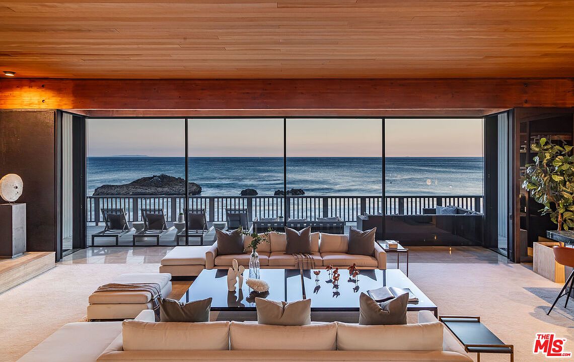 This is a luxurious living room with a stunning ocean view through large, expansive windows. The room features modern, low-profile sofas and a large coffee table, complemented by a warm wood ceiling and neutral-toned flooring. The space is designed for relaxation and entertainment, seamlessly blending indoor and outdoor living.
