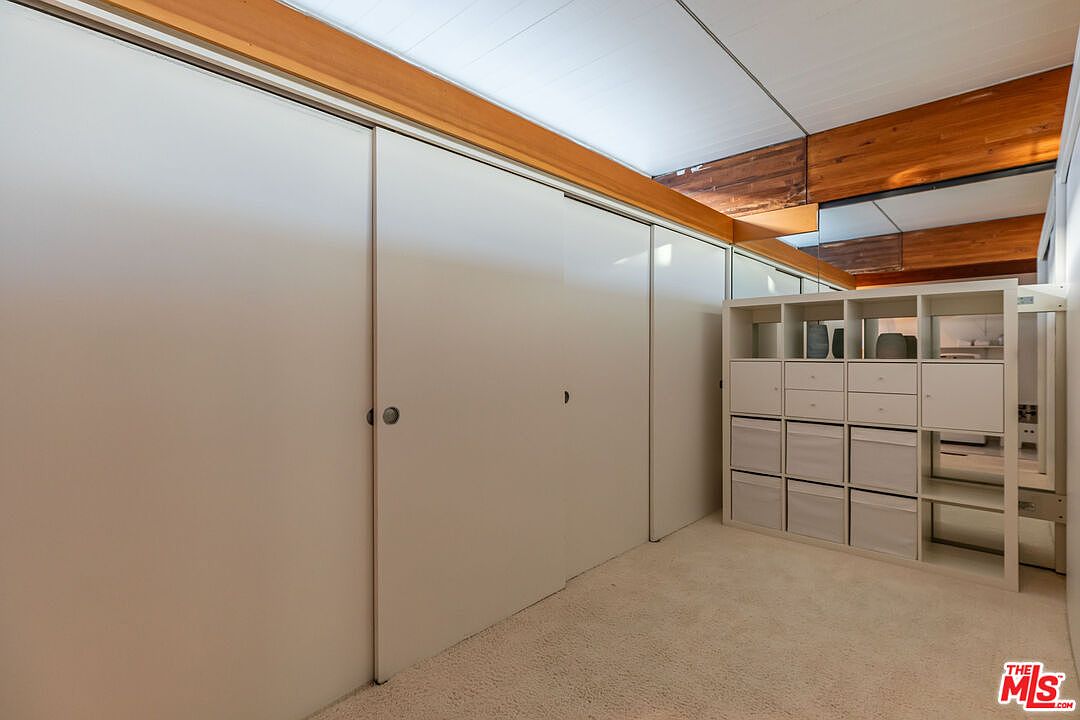 This interior shot showcases a bedroom with a focus on storage solutions. A wall of built-in closets with sliding doors dominates the left side, complemented by a white shelving unit with drawers and compartments on the right. The room is carpeted, and the ceiling features wooden beams, adding a touch of architectural interest.