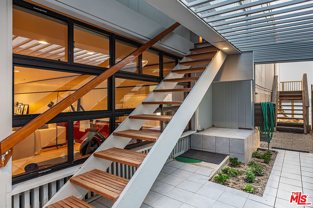 This image showcases an exterior patio area with a unique staircase leading to an upper level. The staircase features wooden steps and a white support structure, complemented by a wooden handrail. The patio is tiled and includes a small garden area, creating a blend of architectural design and natural elements.