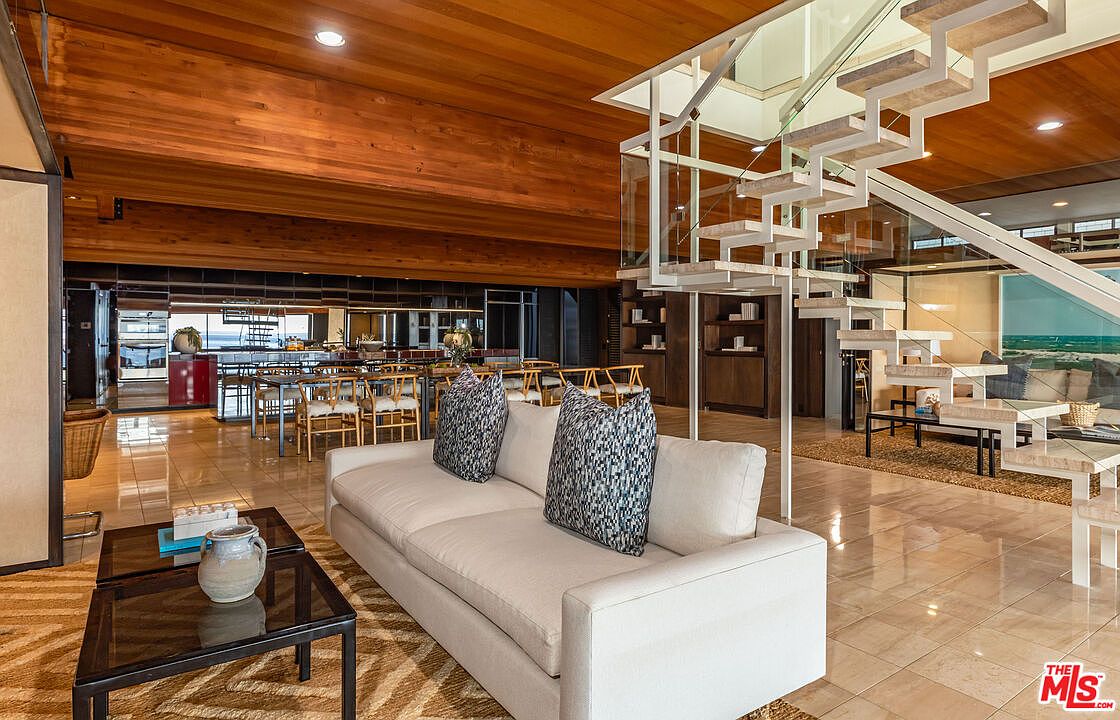This interior shot showcases a spacious living room with a modern design. The room features a light-colored sofa with patterned pillows, a dark coffee table, and a unique staircase with glass railings. The open floor plan leads to a dining area with views of the ocean, and the warm wood ceiling adds a touch of natural elegance.