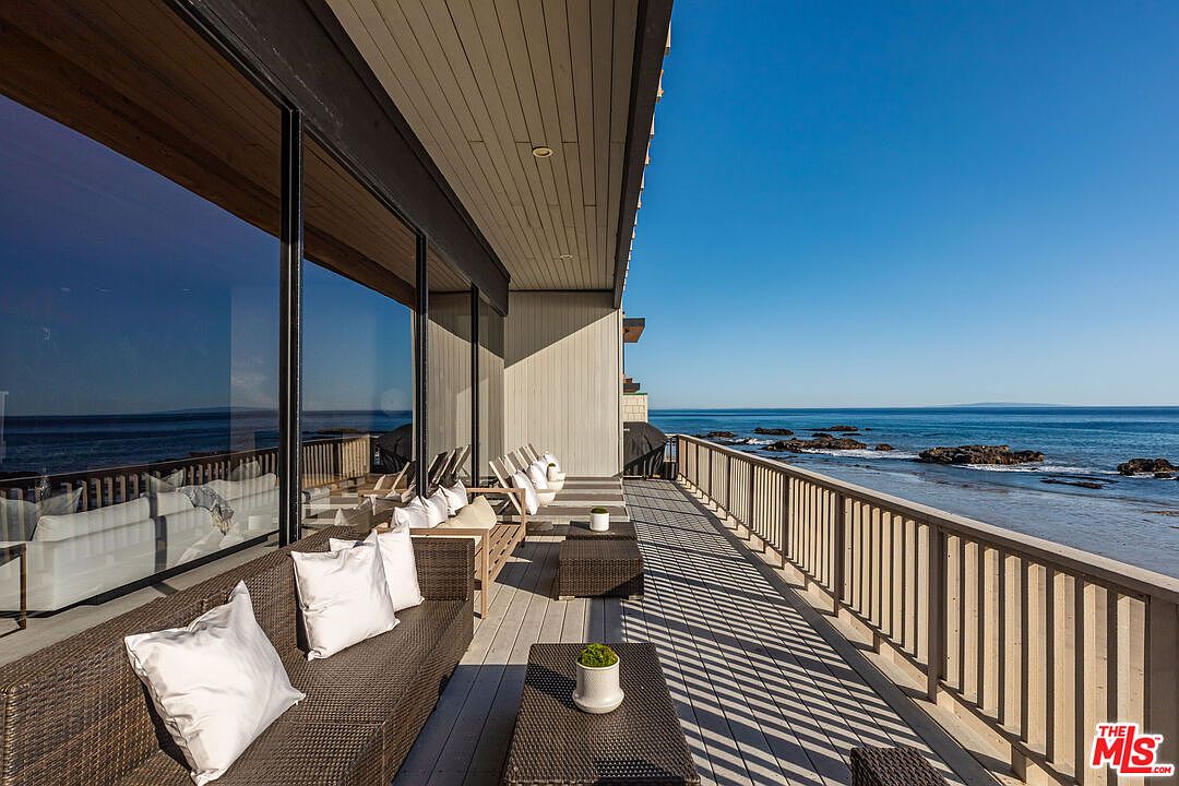 This image showcases a luxurious outdoor patio or deck area with stunning ocean views. The space is furnished with comfortable wicker seating and white cushions, complemented by small tables with potted plants. A wooden railing lines the perimeter, offering safety without obstructing the breathtaking scenery, while large glass windows reflect the ocean view and the interior of the house.