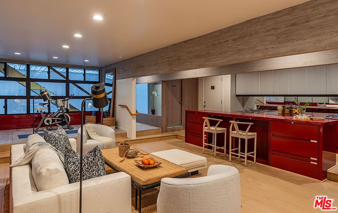 This interior shot showcases a modern living room with a unique design. The room features a seating area with a white sofa and armchairs, a textured coffee table, and a red kitchen island with bar stools. A home gym area is visible in the background, adding to the room's multi-functional appeal.