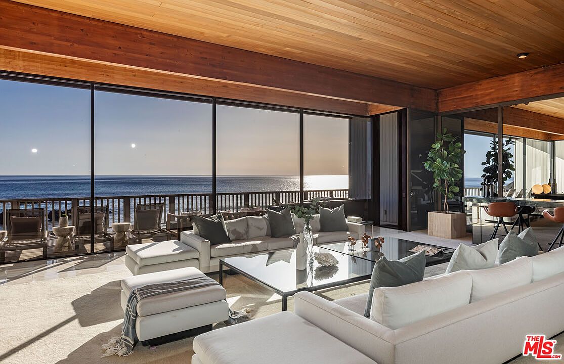 This is a luxurious living room with floor-to-ceiling windows offering stunning ocean views. The room features a modern design with white sofas, a glass coffee table, and a wooden ceiling. The space is bright and airy, creating a relaxing and sophisticated atmosphere.
