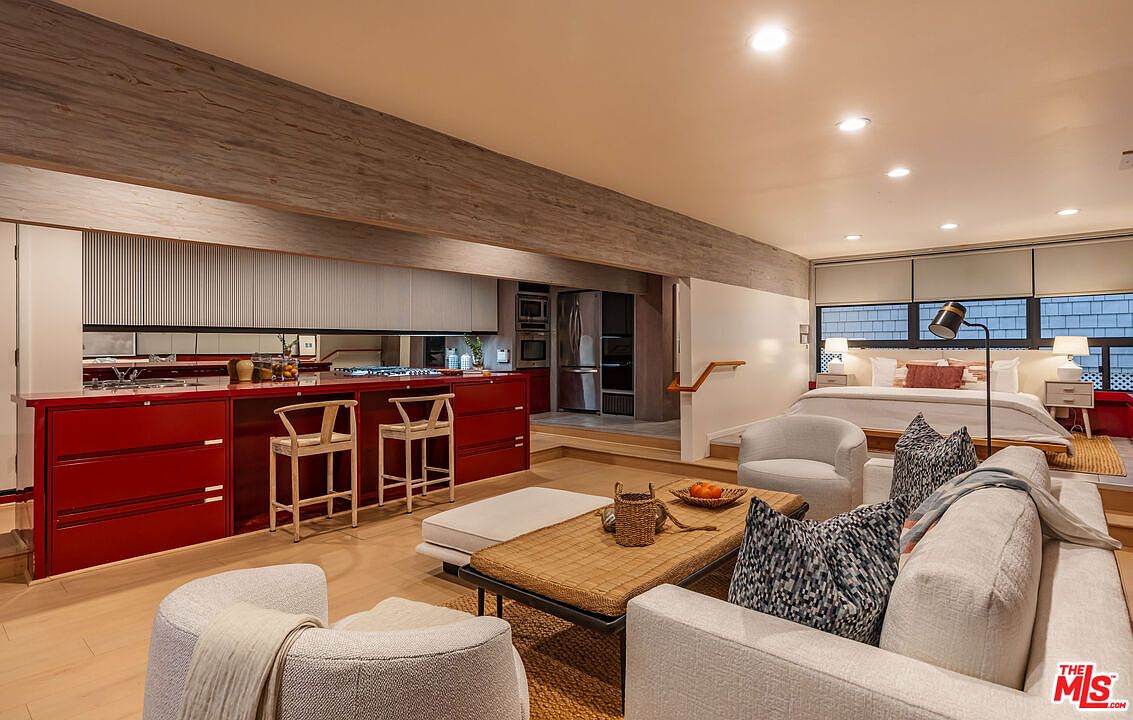 This interior shot showcases a combined living and bedroom space, featuring a modern aesthetic with warm wood flooring and a neutral color palette. The living area includes a light-colored sofa with patterned pillows, a woven coffee table, and comfortable armchairs, while the bedroom area is elevated and features a bed with white linens and bedside tables. The space is well-lit with recessed lighting and natural light filtering through the windows, creating an inviting and comfortable atmosphere.